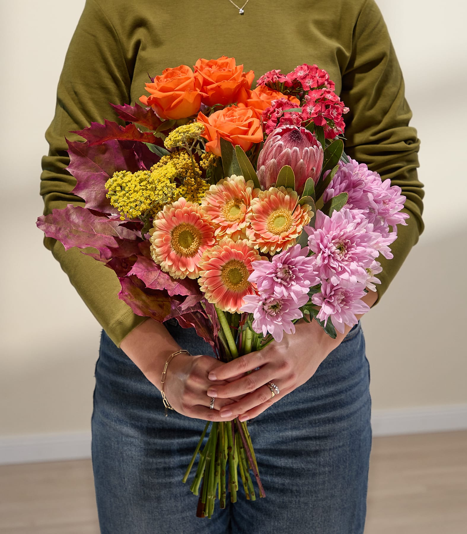 Eine Person in Jeans und einem grünen Pullover hält einen farbenfrohen Blumenstrauß, darunter rosa Chrysanthemen und orangefarbene Rosen, vor einem schlichten, hellen Hintergrund. (Beschreibungstext KI-generiert)