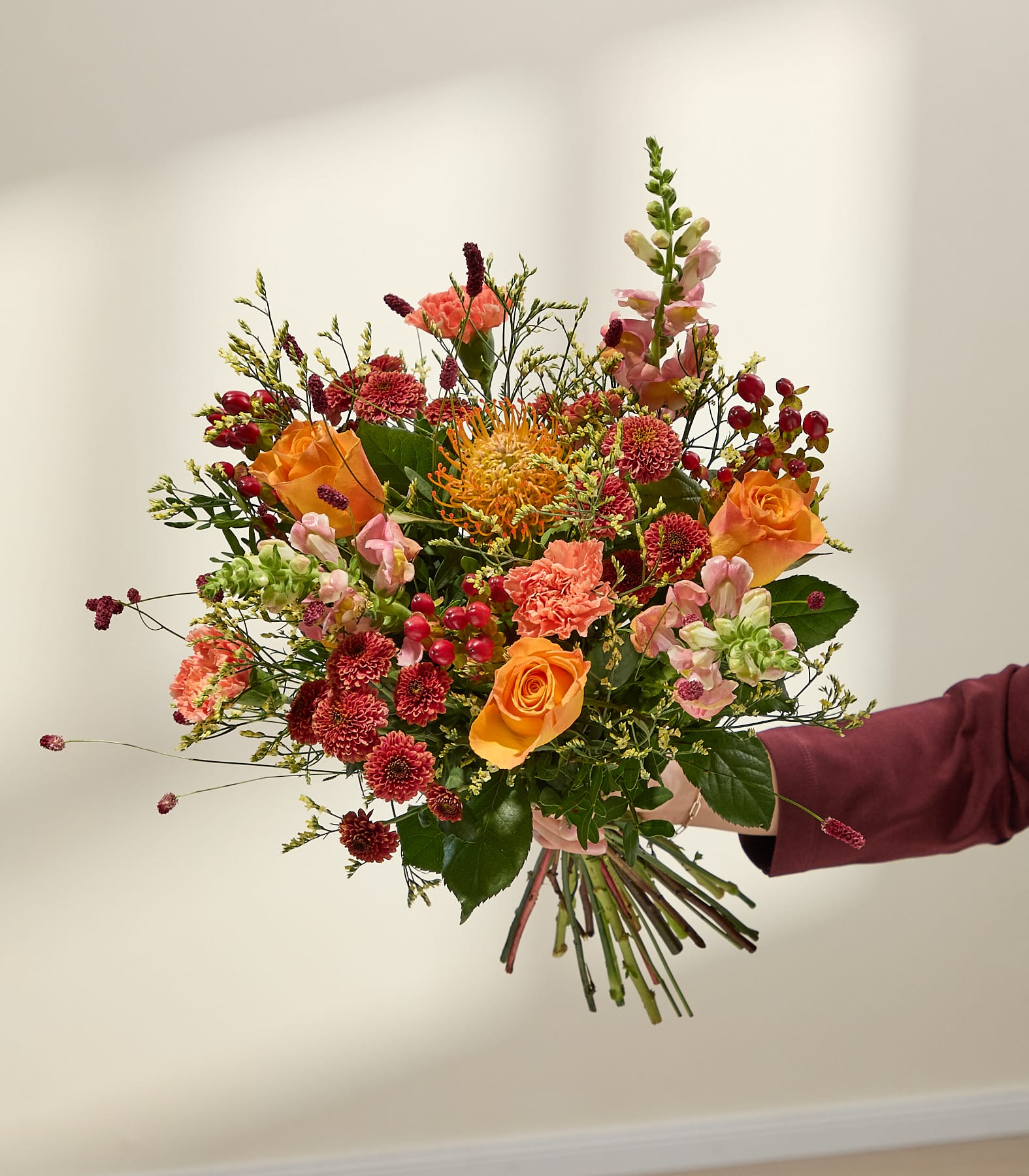Auf einem Fensterbrett steht ein farbenfroher Strauß herbstlicher Blumen in einer strukturierten Glasvase, im Vordergrund dekorative Kerzen und im Hintergrund ein Fenster mit gefiltertem Licht. (Beschreibungstext KI-generiert)