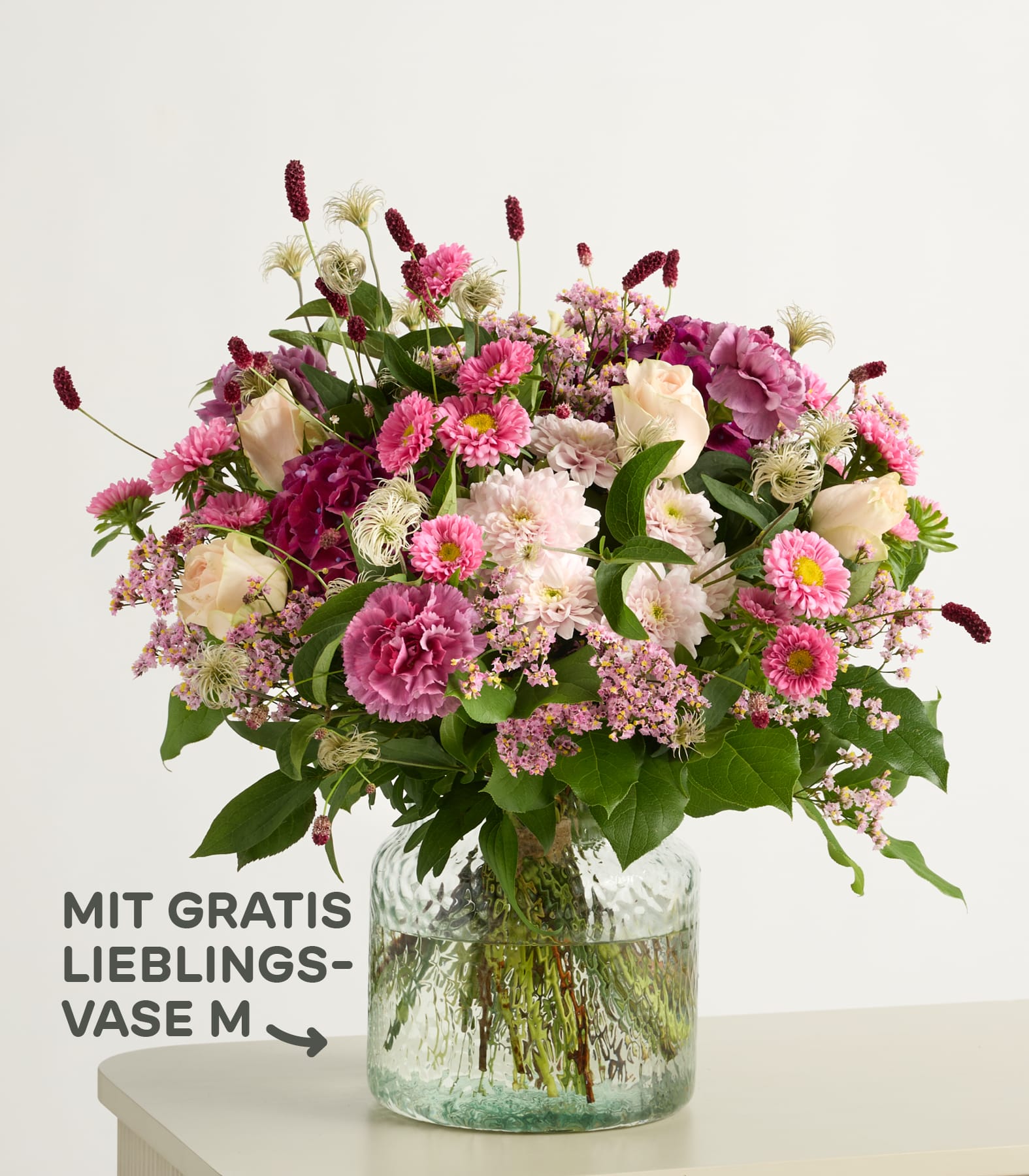 Im Vordergrund füllt ein farbenfroher Strauß rosa und weißer Blumen eine strukturierte Glasvase vor einem schlichten, hellen Hintergrund. (Beschreibungstext KI-generiert)