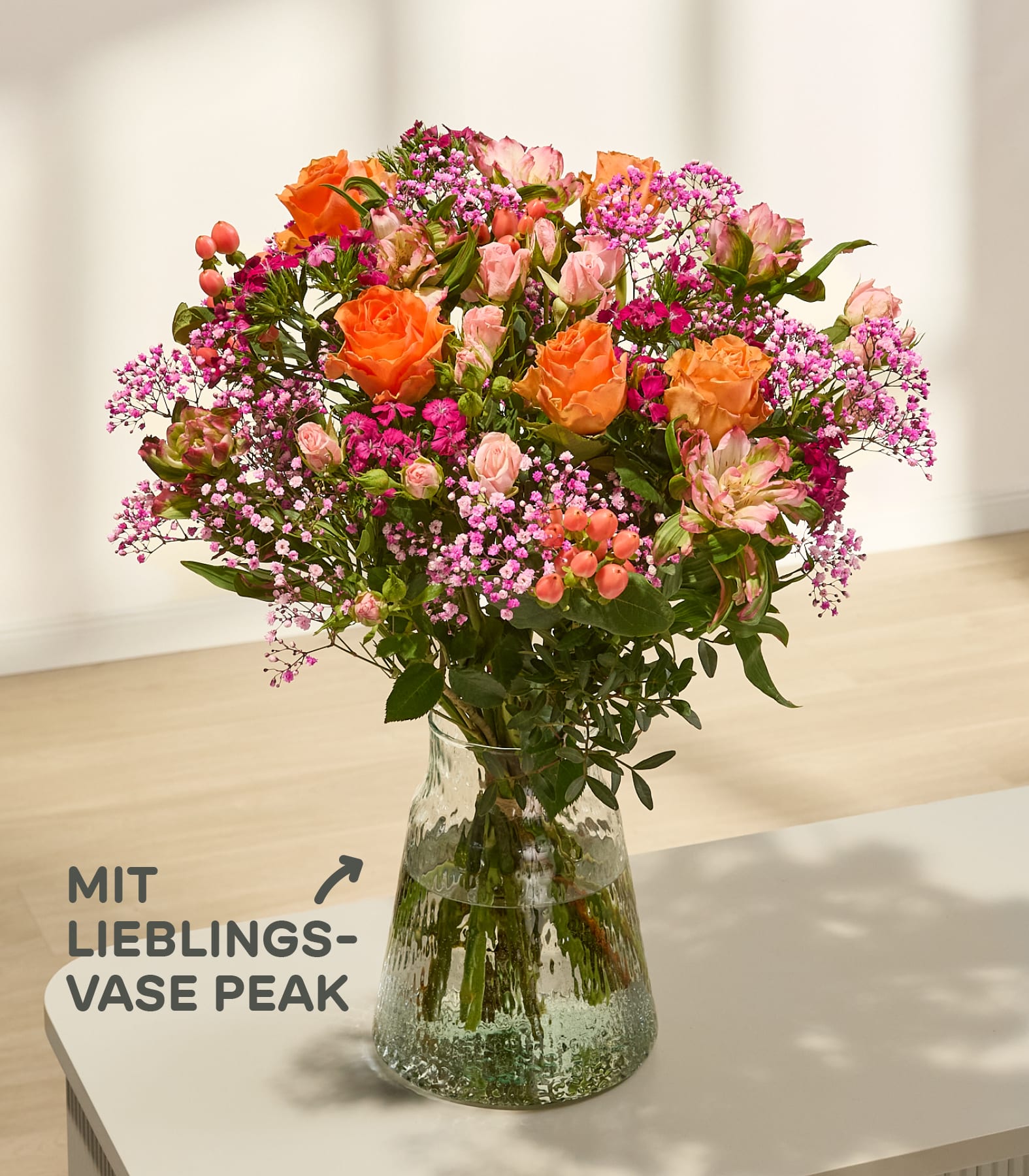 Ein leuchtender Strauß aus orangefarbenen und rosafarbenen Rosen mit zarten rosafarbenen Füllblumen steht in einer strukturierten Glasvase auf einer weißen Fläche, im Hintergrund sind Wand und Boden verschwommen zu sehen. (Beschreibungstext KI-generiert)