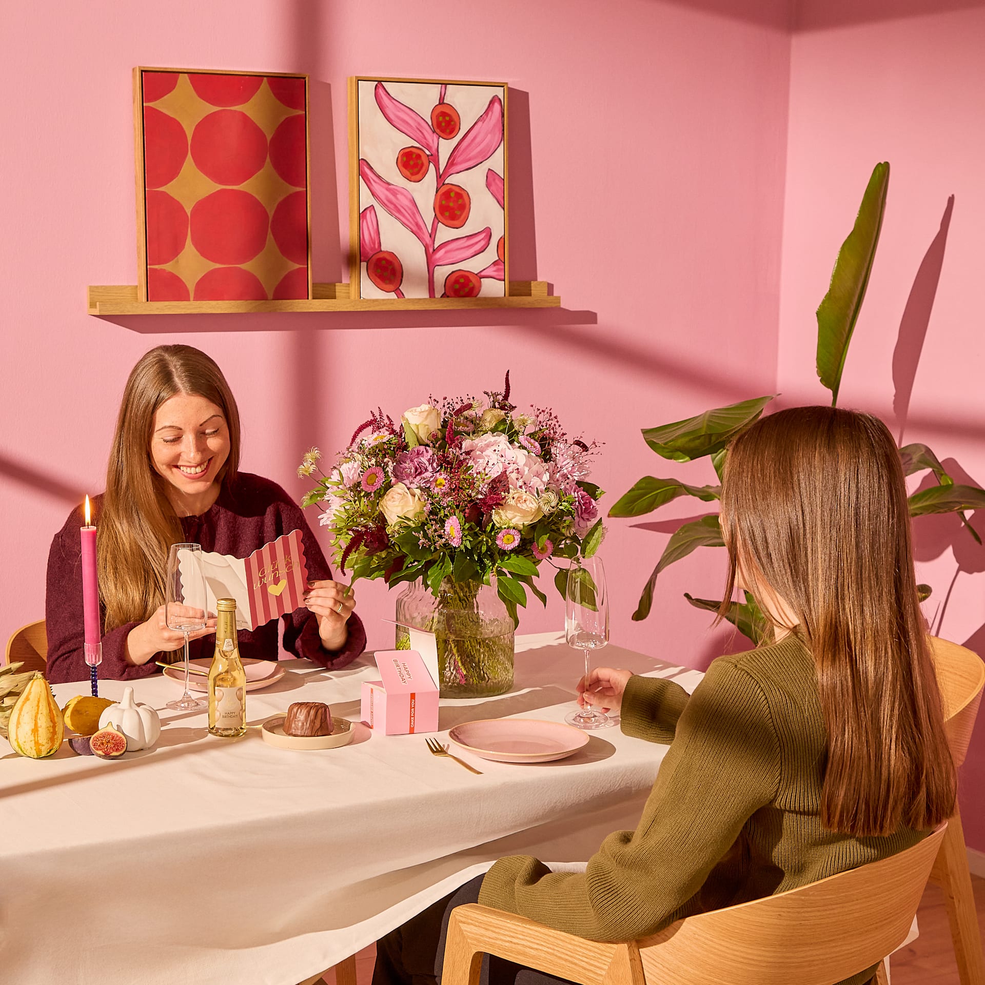 Zwei Frauen sitzen an einem mit Blumenarrangements und Desserts gedeckten Tisch; im Hintergrund sind rosa Wände und Kunstwerke zu sehen. (Beschreibungstext KI-generiert)