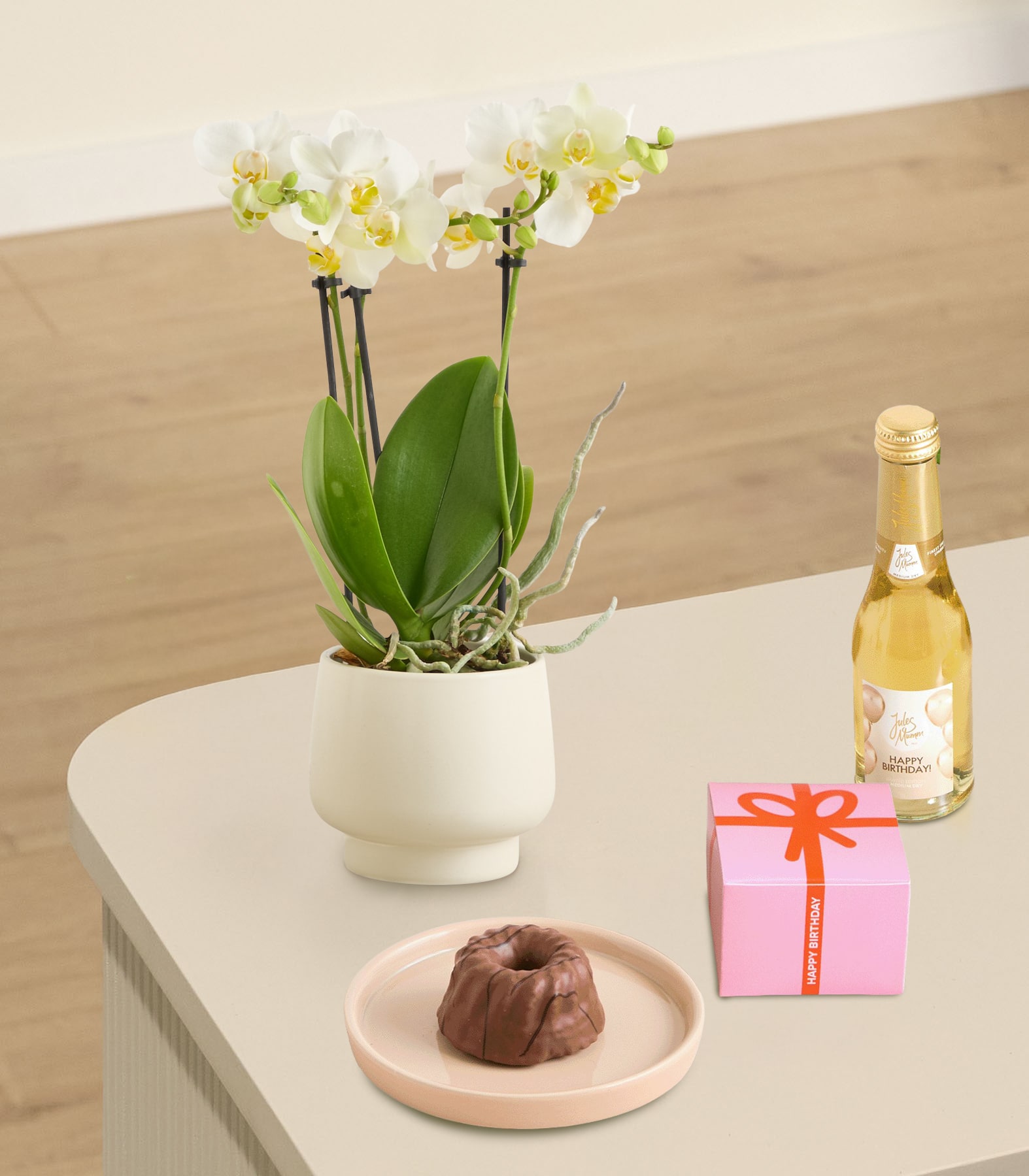 Auf einer Holzoberfläche steht eine Orchidee im Topf mit zarten gelben Blüten, daneben eine kleine Geschenkbox und eine Flasche, die Champagner oder Sekt zu sein scheint. (Beschreibungstext KI-generiert)