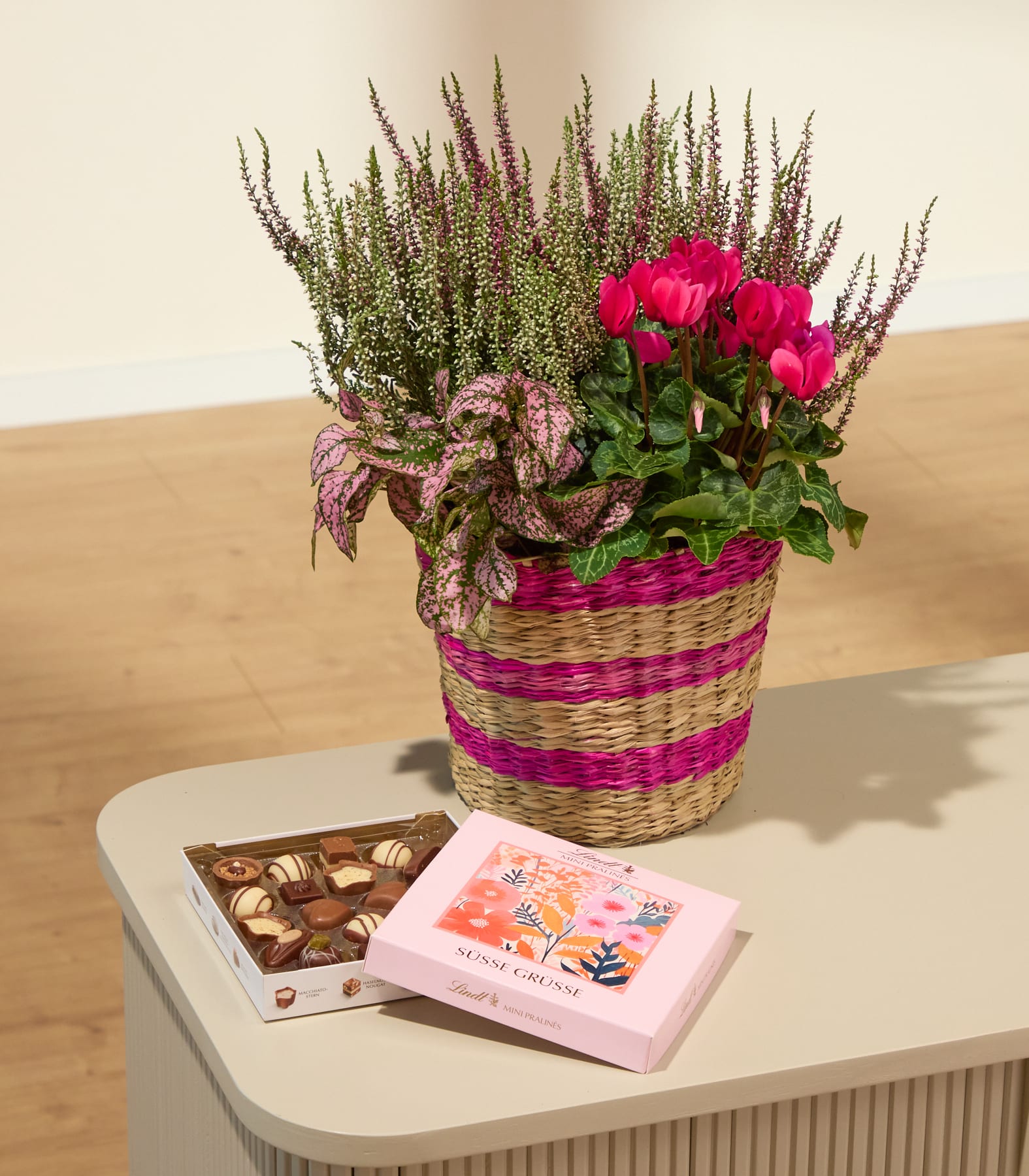 Ein geflochtener Korb, gefüllt mit rosa und violetten Blütenpflanzen, steht hinter einer Schachtel Pralinen und einer rosa Blumenbox auf einem hellen Tisch; im Hintergrund sind ein Holzboden und eine cremefarbene Wand zu sehen. (Beschreibungstext KI-generiert)