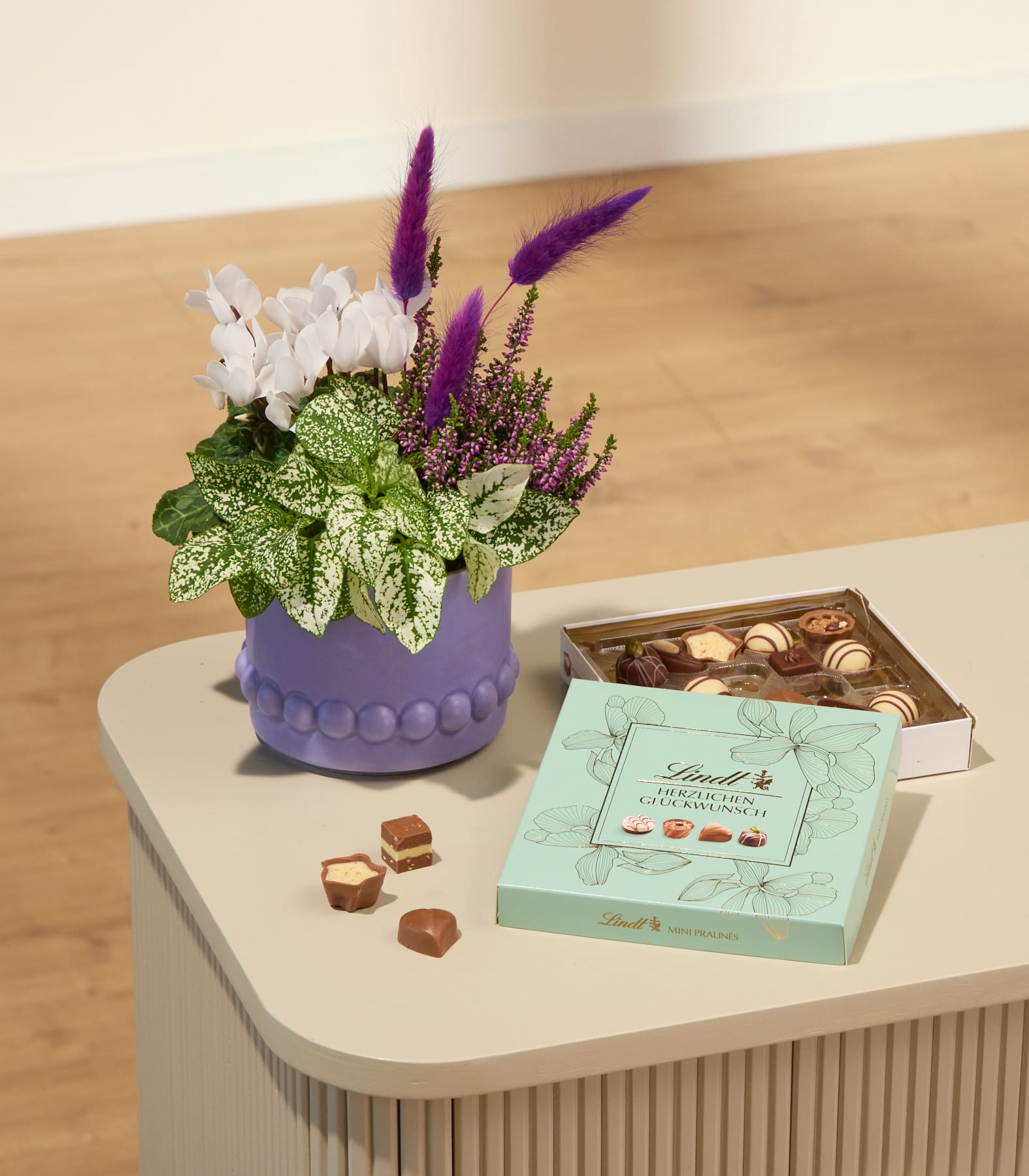 Auf einem hellen Tisch steht ein violetter Topf mit einer blühenden Pflanze und violetten Akzenten neben zwei Schachteln Pralinen und einigen verstreuten Pralinen; im Hintergrund sind ein verschwommener Holzboden und eine Holzwand zu sehen. (Beschreibungstext KI-generiert)