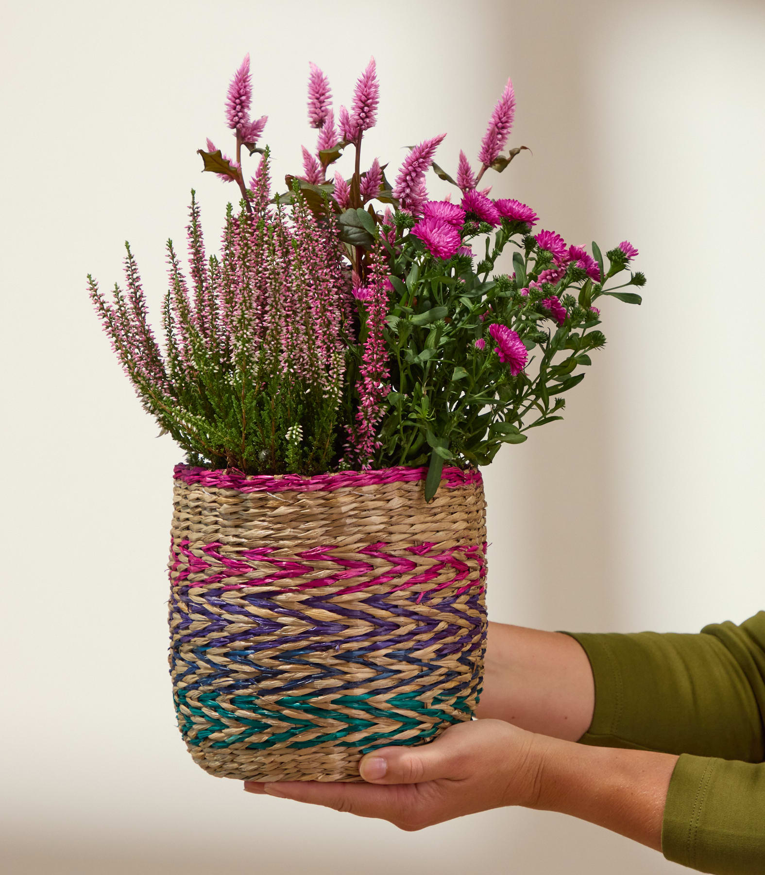 Zwei Hände halten einen geflochtenen Korb, gefüllt mit rosa Heidekraut und Chrysanthemen, vor einem schlichten Hintergrund. (Beschreibungstext KI-generiert)
