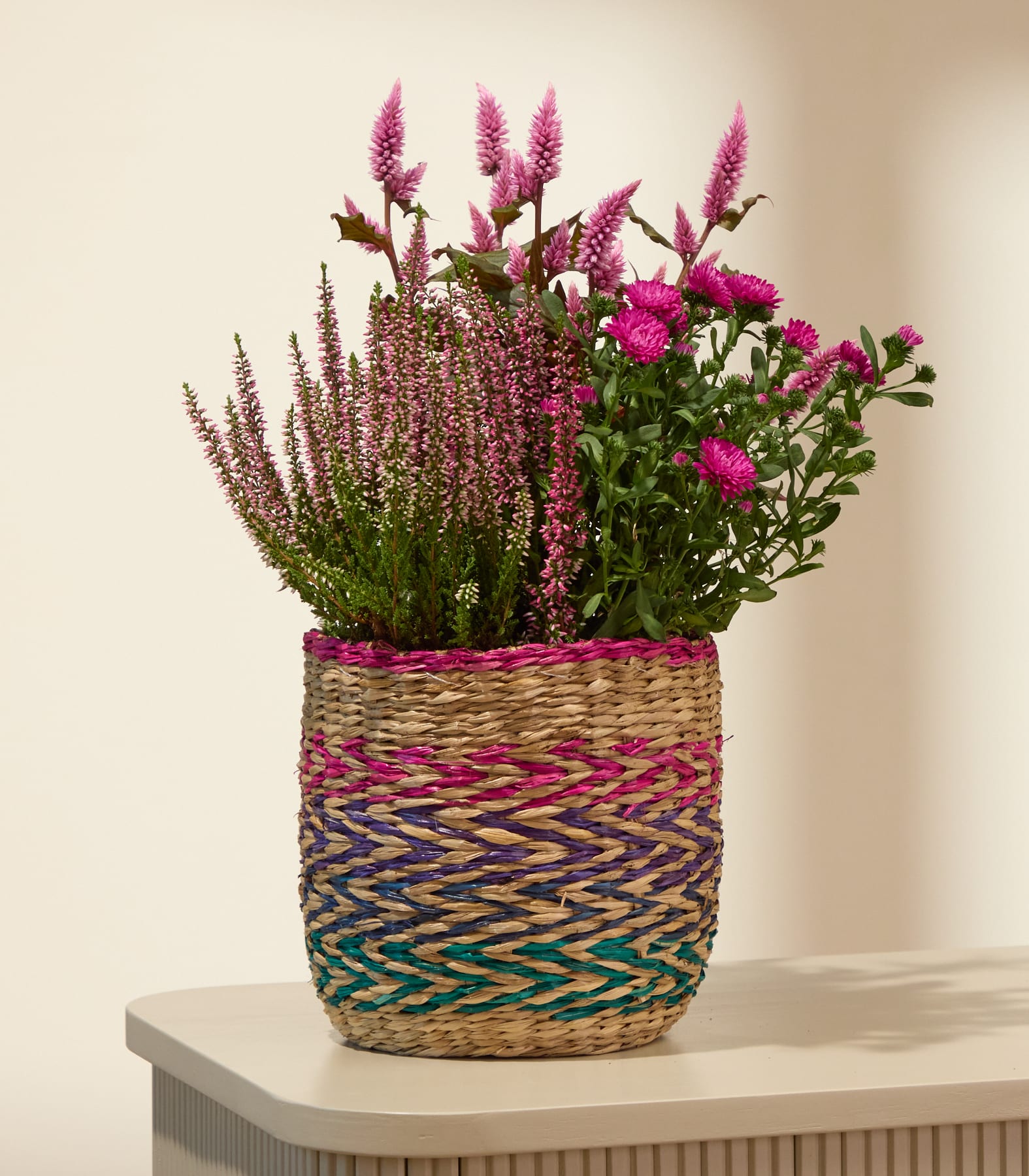 Ein geflochtener Korb, gefüllt mit rosa Heidekraut und Chrysanthemen, steht auf einer hellen Oberfläche vor einem sanften, neutralen Hintergrund. (Beschreibungstext KI-generiert)