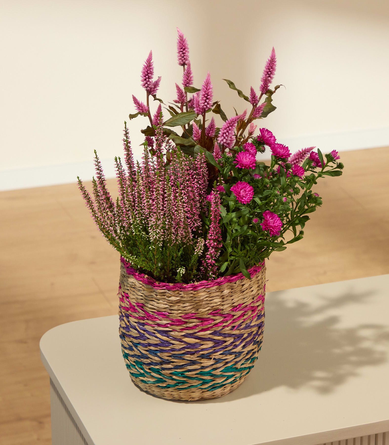 Ein geflochtener Korb, gefüllt mit rosa Blumen, steht auf einer hellen Fläche; im Hintergrund sind Wand und Boden verschwommen zu erkennen. (Beschreibungstext KI-generiert)