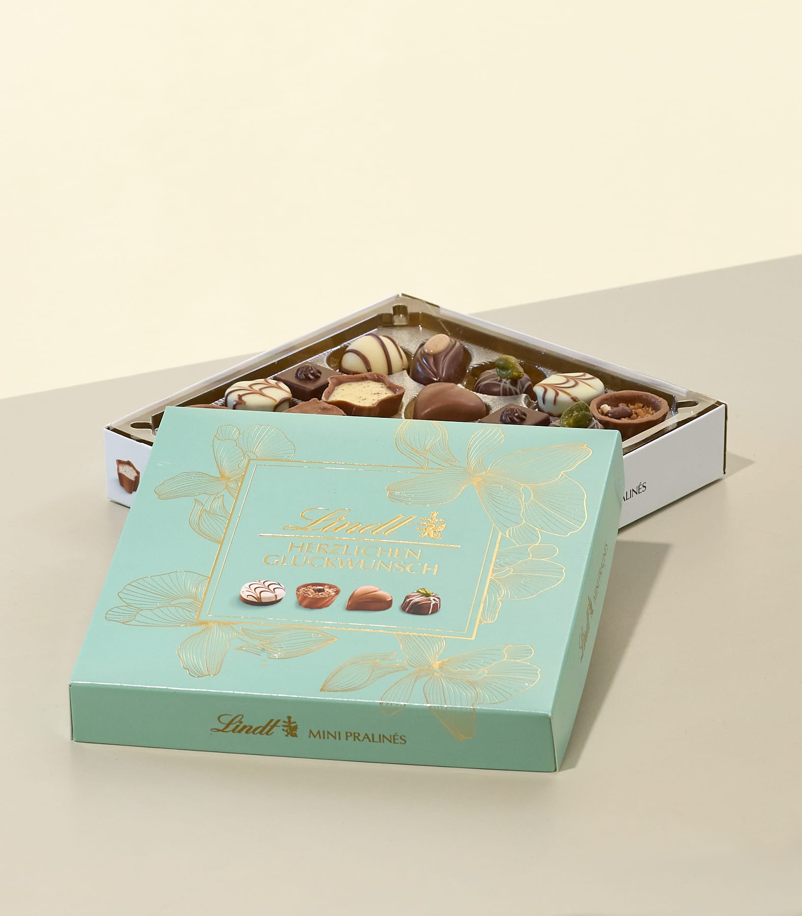Im Vordergrund steht eine helltürkisfarbene Lindt-Schachtel mit goldenen Blumenverzierungen geöffnet und gibt den Blick auf eine Auswahl an Mini-Pralinen frei, während im Hintergrund vor einer hellen Wand eine zweite, teilweise geöffnete Schachtel mit verschiedenen Pralinen zu sehen ist. (Beschreibungstext KI-generiert)