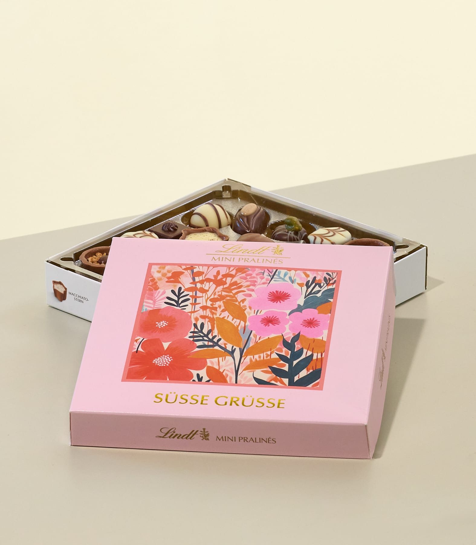 Im Vordergrund steht eine rosafarbene Schachtel Lindt Mini-Pralinen mit Blumenmuster, während im Hintergrund vor einer hellbeigen Wand eine offene Schachtel mit Pralinenmischung zu sehen ist. (Beschreibungstext KI-generiert)
