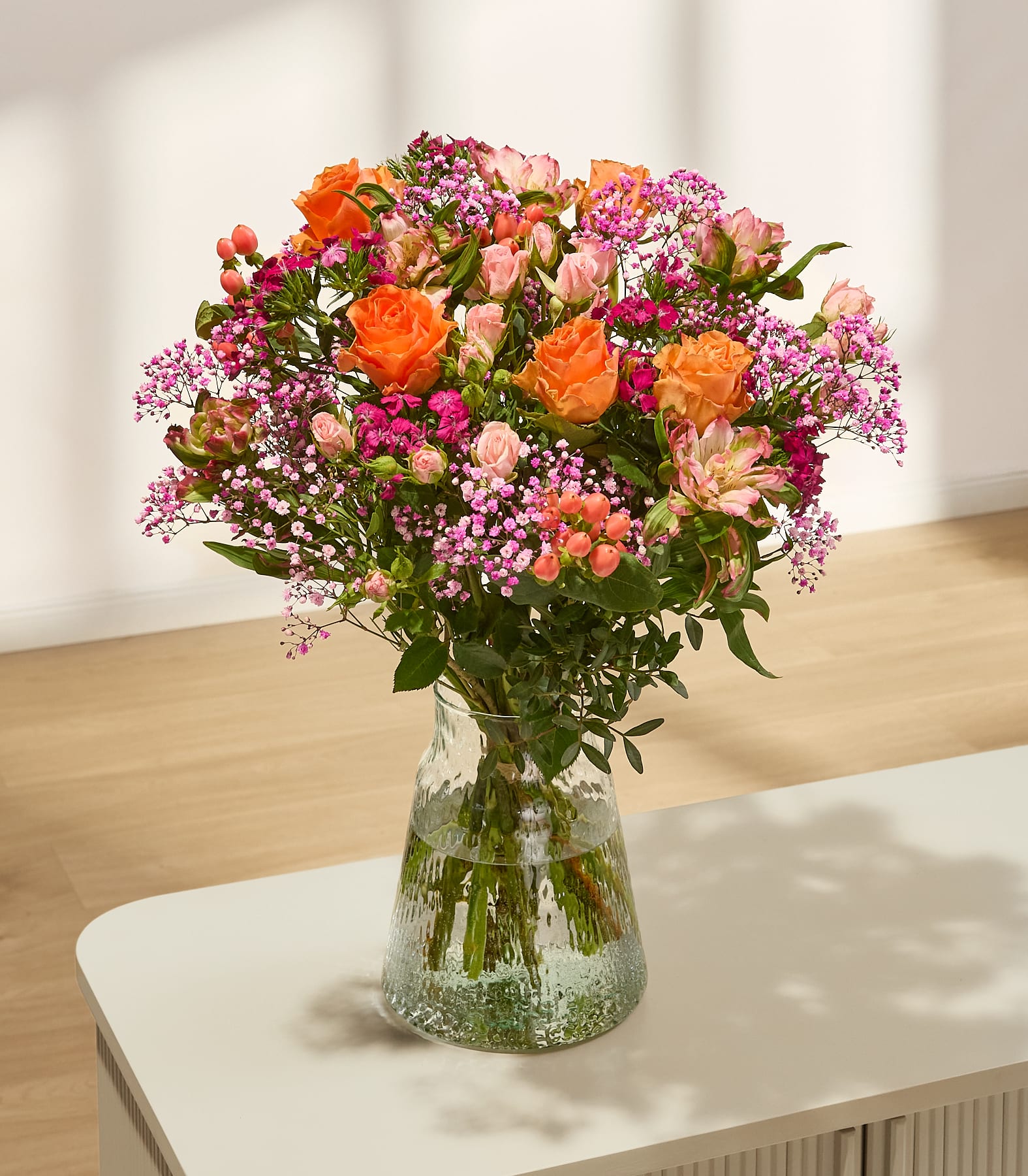 Ein lebhafter Strauß bunter Blumen, darunter rosa und gelbe Rosen, weiße Gänseblümchen und andere Blüten, in den Händen einer Person vor einem schlichten, hellen Hintergrund. (Beschreibungstext KI-generiert)