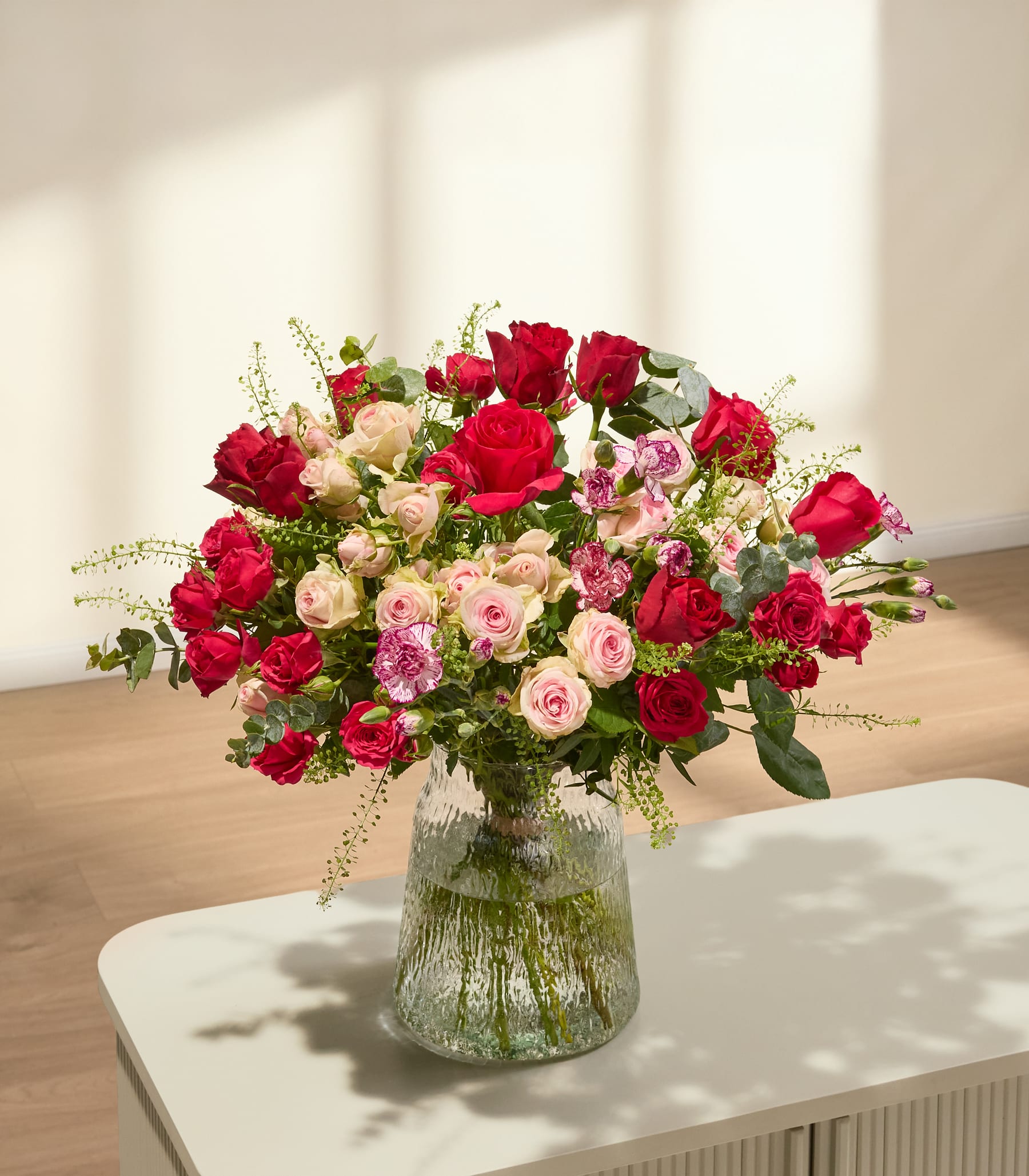 Ein großer, farbenprächtiger Strauß roter und rosa Rosen, Nelken und anderer Blumen, der von einer Person in den Händen gehalten wird, vor einem schlichten weißen Hintergrund. (Beschreibungstext KI-generiert)