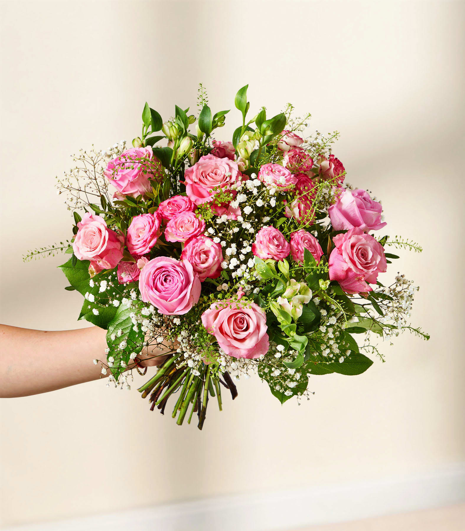 Eine Hand hält einen üppigen Strauß rosa Rosen und Schleierkraut vor einem sanft verschwommenen Hintergrund aus hellen Wänden und einer Holzoberfläche. (Beschreibungstext KI-generiert)