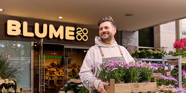 Ein Mann mit Bart und in Freizeitkleidung steht vor einem Blumenladen namens „Blume 2000“ und hält eine Kiste mit bunten Blumen.