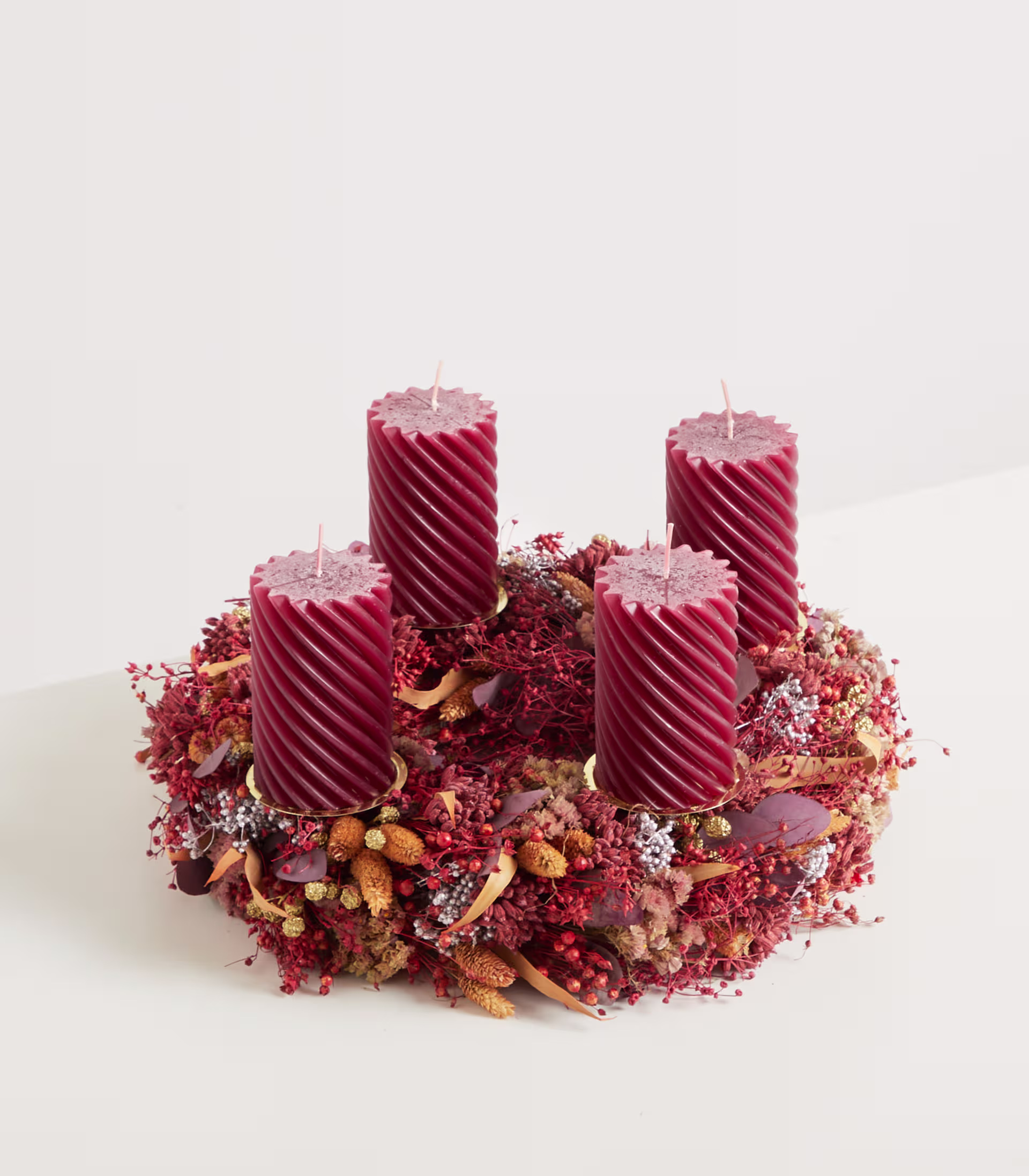 Das Bild zeigt ein festliches kranzartiges Arrangement mit vier roten Spiralkerzen, umgeben von einer Mischung aus getrockneten Blumen, Beeren und anderen natürlichen Elementen auf einem schlichten weißen Hintergrund. (Beschreibungstext KI-generiert)
