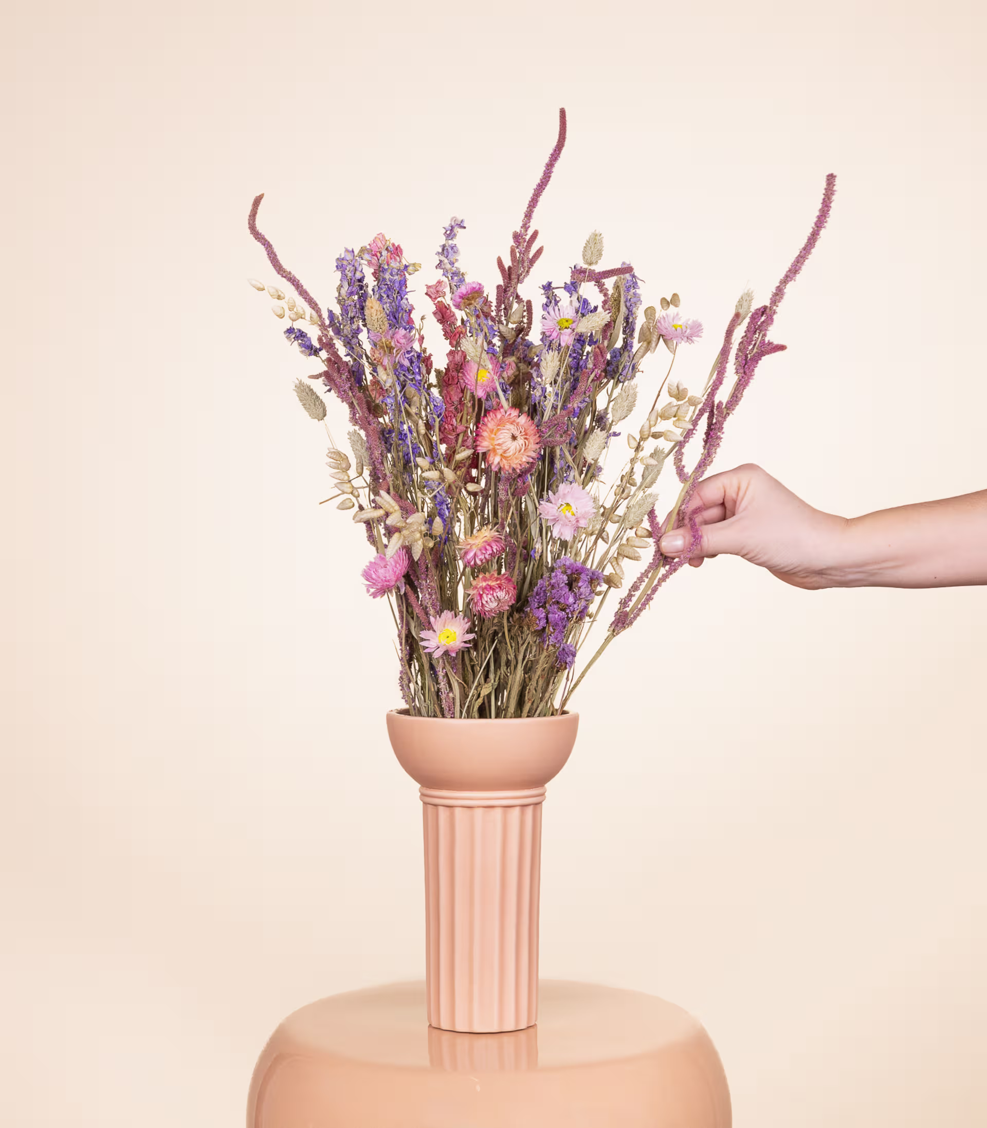Eine Hand hält einen Strauß getrockneter Blumen in einer Keramikvase vor einem schlichten beigen Hintergrund. (Beschreibungstext KI-generiert)