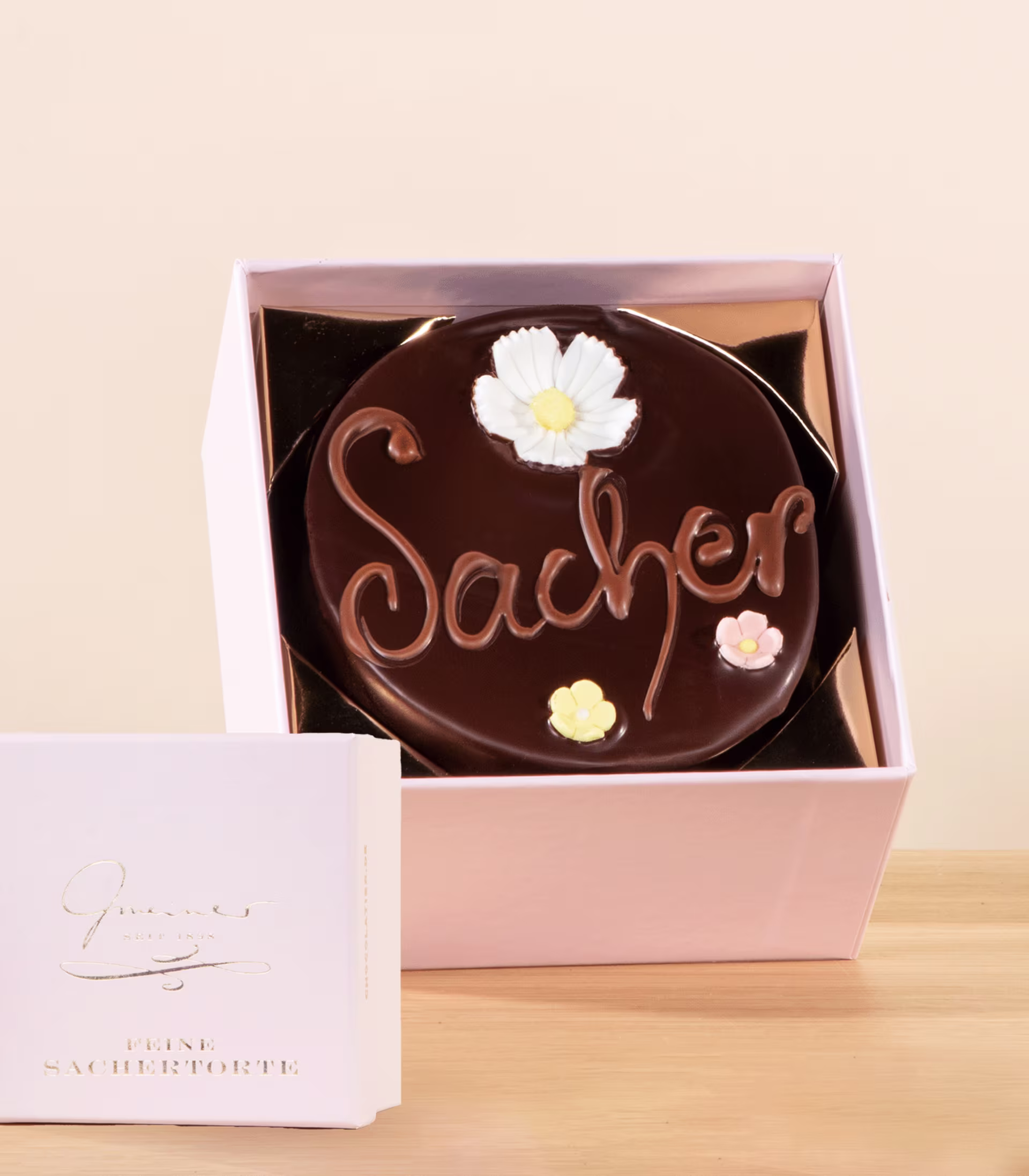 Eine Schokoladentorte mit der Aufschrift „Sacher“, dekoriert mit weißen Blumen, steht in einer rosa Schachtel auf einer Holzoberfläche. (Beschreibungstext KI-generiert)