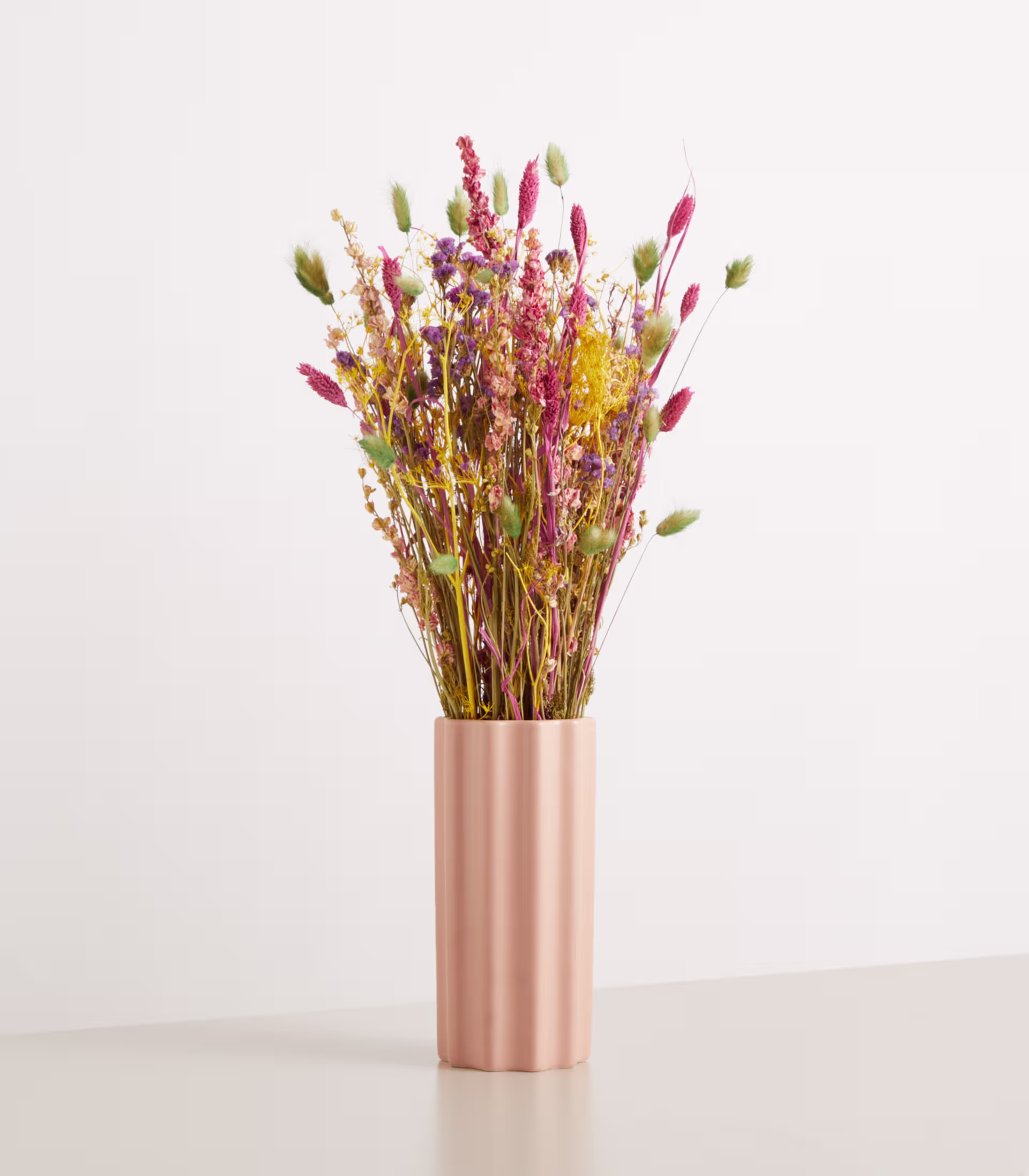 Ein Strauß leuchtender Trockenblumen in einer metallisch rosa Vase steht vor einem schlichten weißen Hintergrund. (Beschreibungstext KI-generiert)