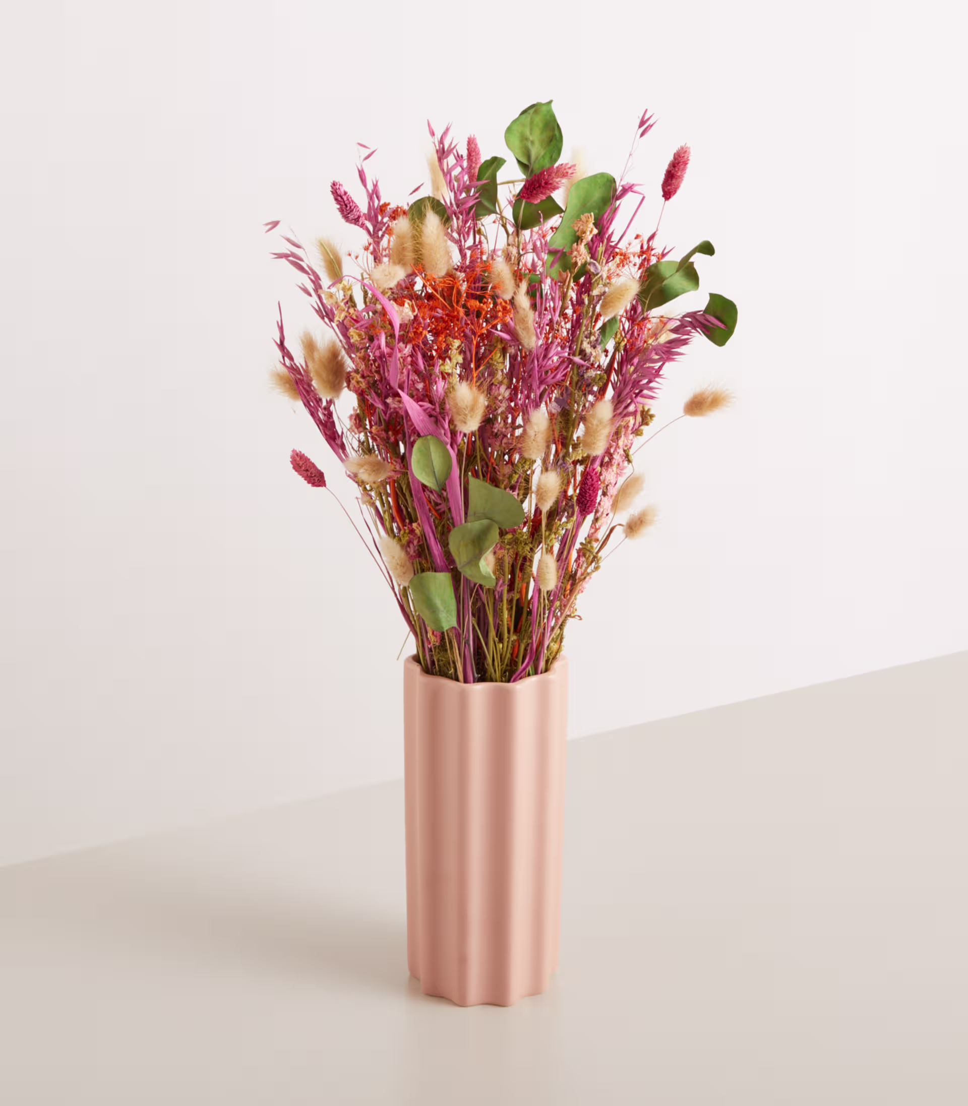Ein farbenfroher Strauß getrockneter Blumen in einer metallisch rosa Vase steht vor einem schlichten, hellen Hintergrund. (Beschreibungstext KI-generiert)