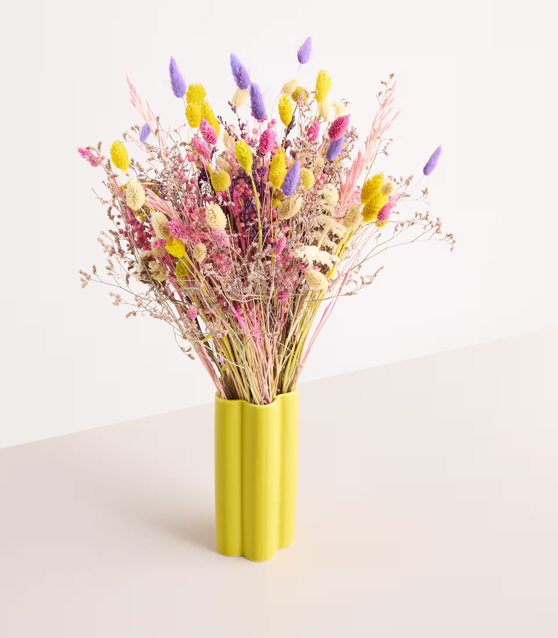 Ein farbenfroher Strauß getrockneter Blumen in verschiedenen Lila-, Gelb- und Rosatönen wird in einer leuchtend gelben Vase vor einem schlichten weißen Hintergrund präsentiert. (Beschreibungstext KI-generiert)