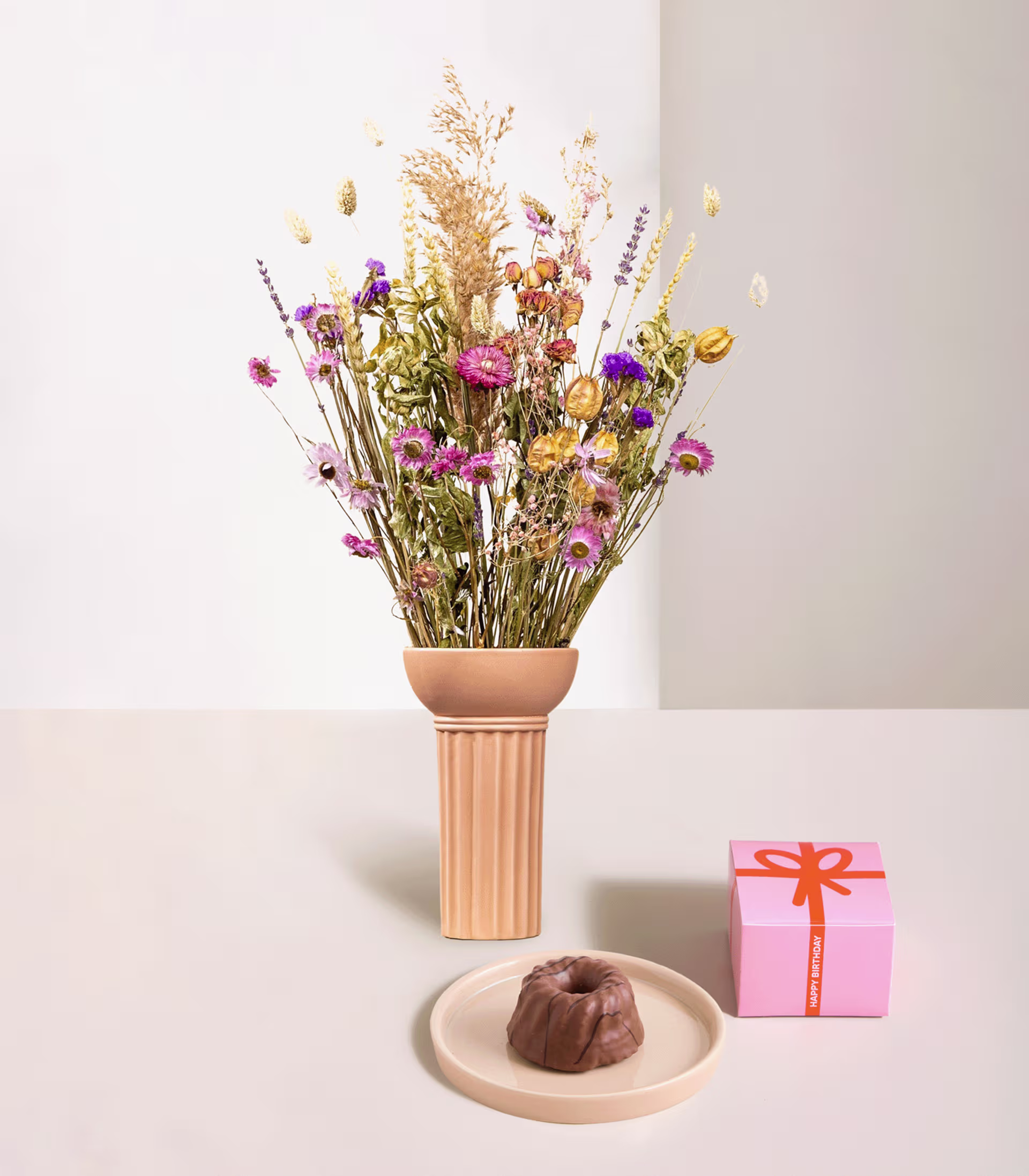 Ein farbenfroher Strauß getrockneter Blumen steht in einer Keramikvase auf einem Tisch, begleitet von einer kleinen rosa Geschenkbox und einem Schokoladenkonfekt auf einem Teller. (Beschreibungstext KI-generiert)