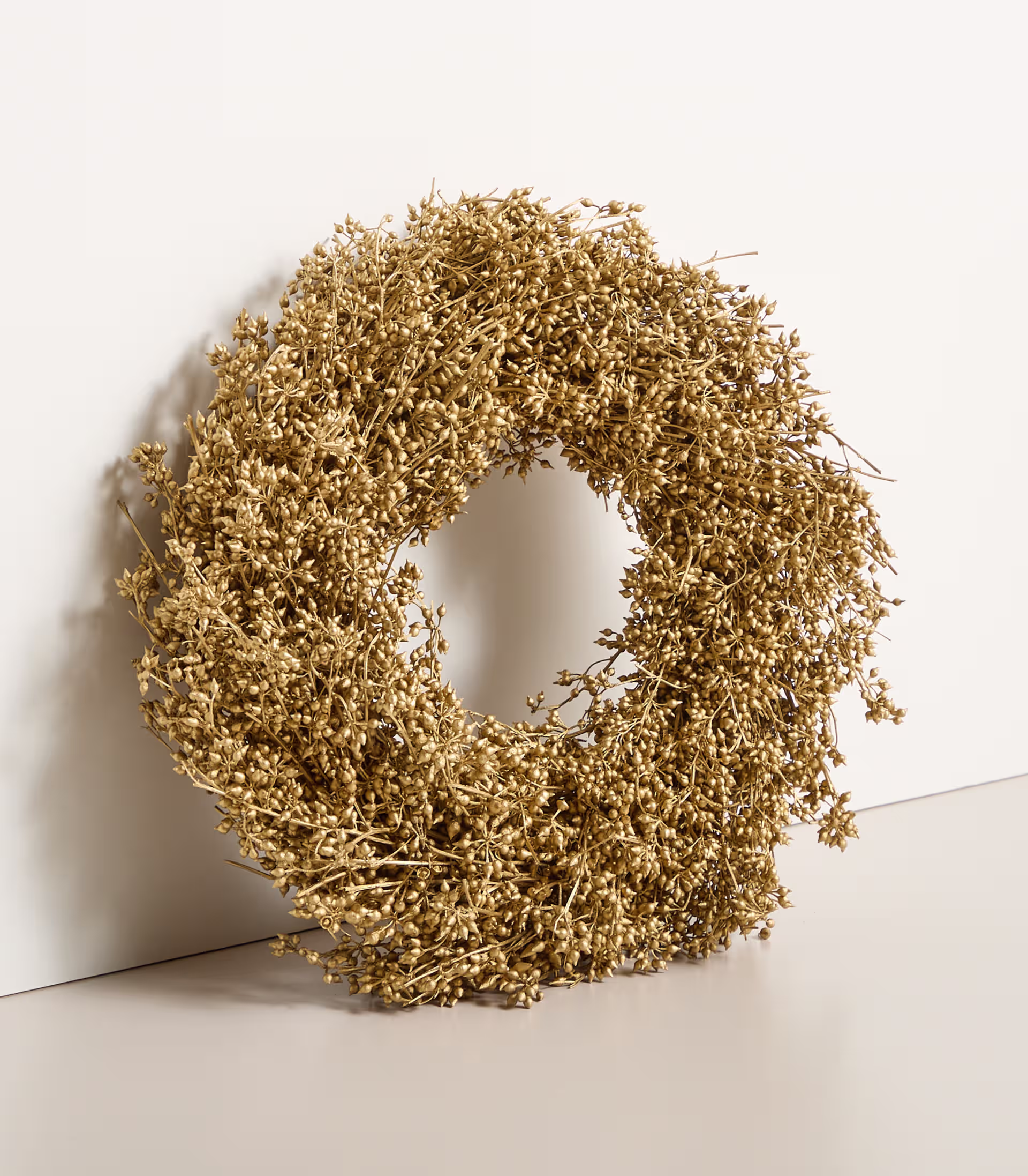 Auf einem schlichten weißen Hintergrund befindet sich ein goldener Kranz aus getrockneten Blumen oder Blättern. (Beschreibungstext KI-generiert)