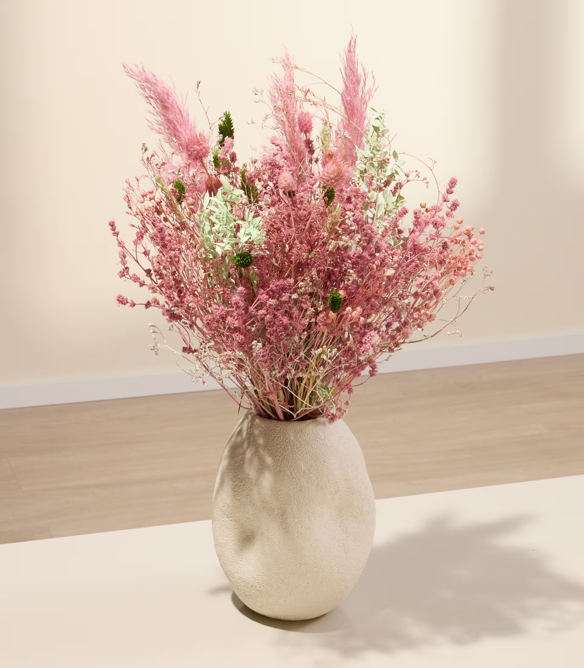 Eine Vase, gefüllt mit einem farbenfrohen Strauß aus rosa und weißen Trockenblumen, steht auf einer Holzoberfläche vor einem schlichten Hintergrund. (Beschreibungstext KI-generiert)