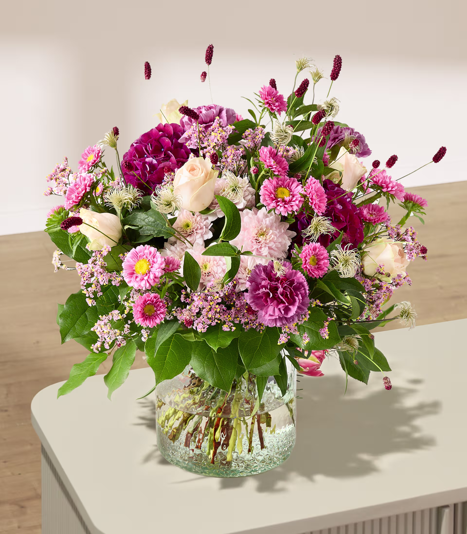 Ein leuchtender Strauß rosa und weißer Blumen füllt eine durchsichtige Glasvase auf hellem Untergrund vor einem weichen, verschwommenen Hintergrund. (Beschreibungstext KI-generiert)