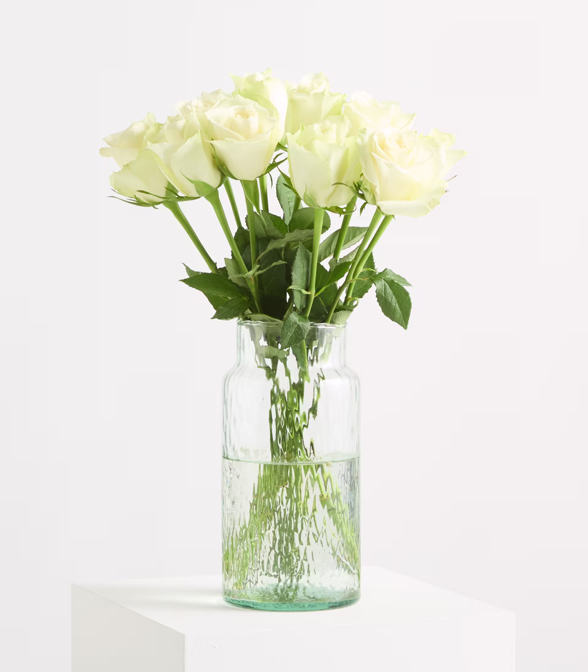 Ein Strauß weißer Rosen in einer strukturierten Glasvase steht auf einem weißen Sockel vor einem schlichten weißen Hintergrund. (Beschreibungstext KI-generiert)