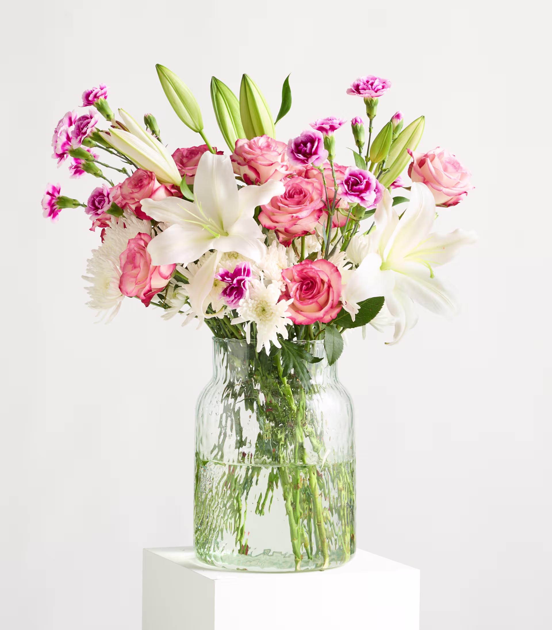 Ein leuchtender Strauß aus rosa Rosen, weißen Lilien und Nelken steht in einer durchsichtigen Glasvase auf einem weißen Sockel vor einem schlichten weißen Hintergrund. (Beschreibungstext KI-generiert)