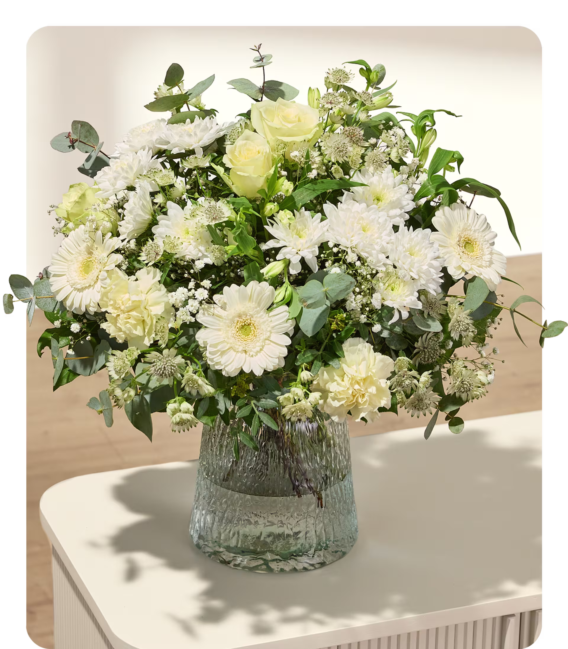Ein üppiger Strauß weißer und cremefarbener Blumen in einer strukturierten Glasvase steht auf einem weißen Tisch vor einem schlichten, hellen Hintergrund. (Beschreibungstext KI-generiert)