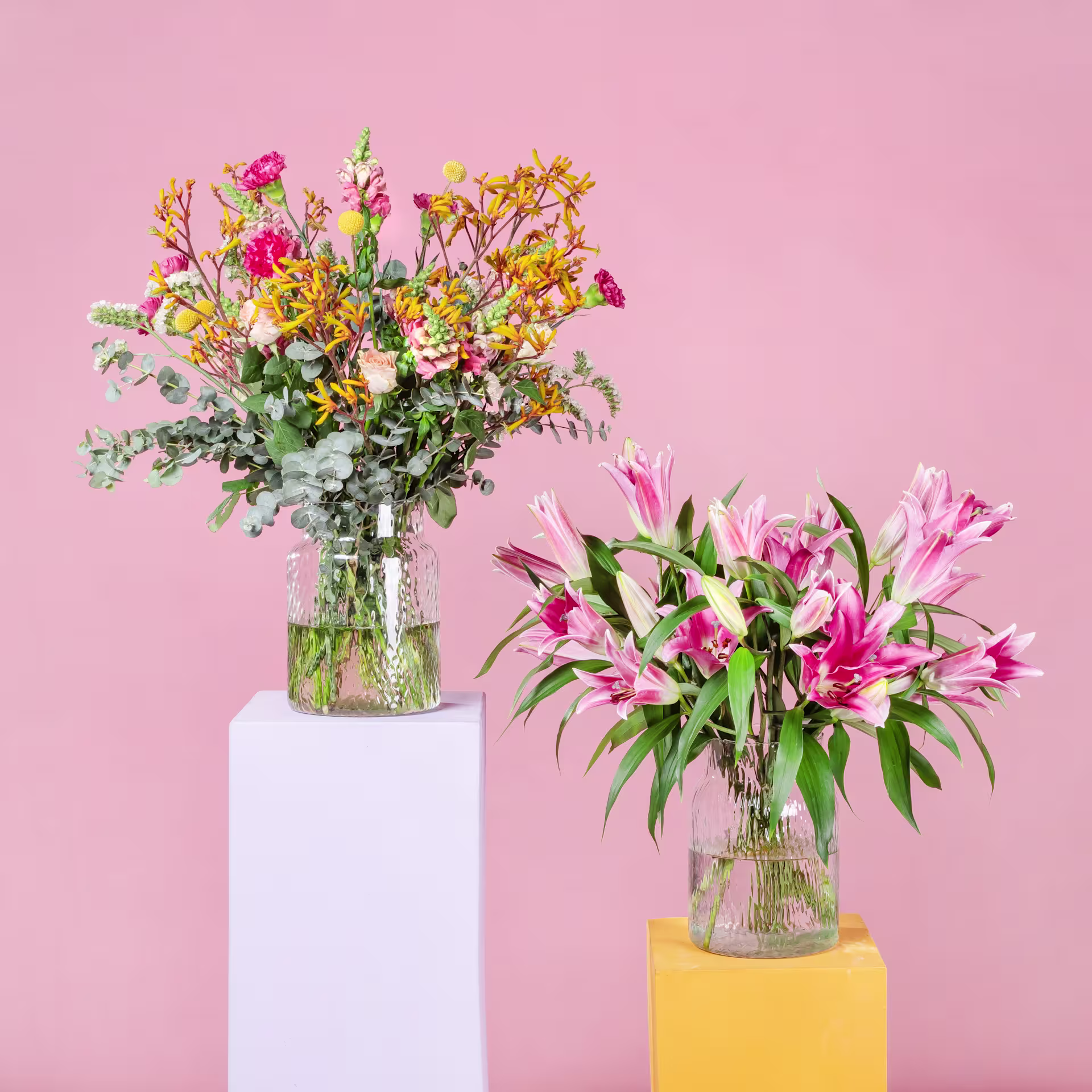 Ein leuchtender Strauß bunter Blumen, darunter rosa Lilien und gelbe Wildblumen, steht auf einem weißen Sockel vor einem zartrosa Hintergrund. (Beschreibungstext KI-generiert)