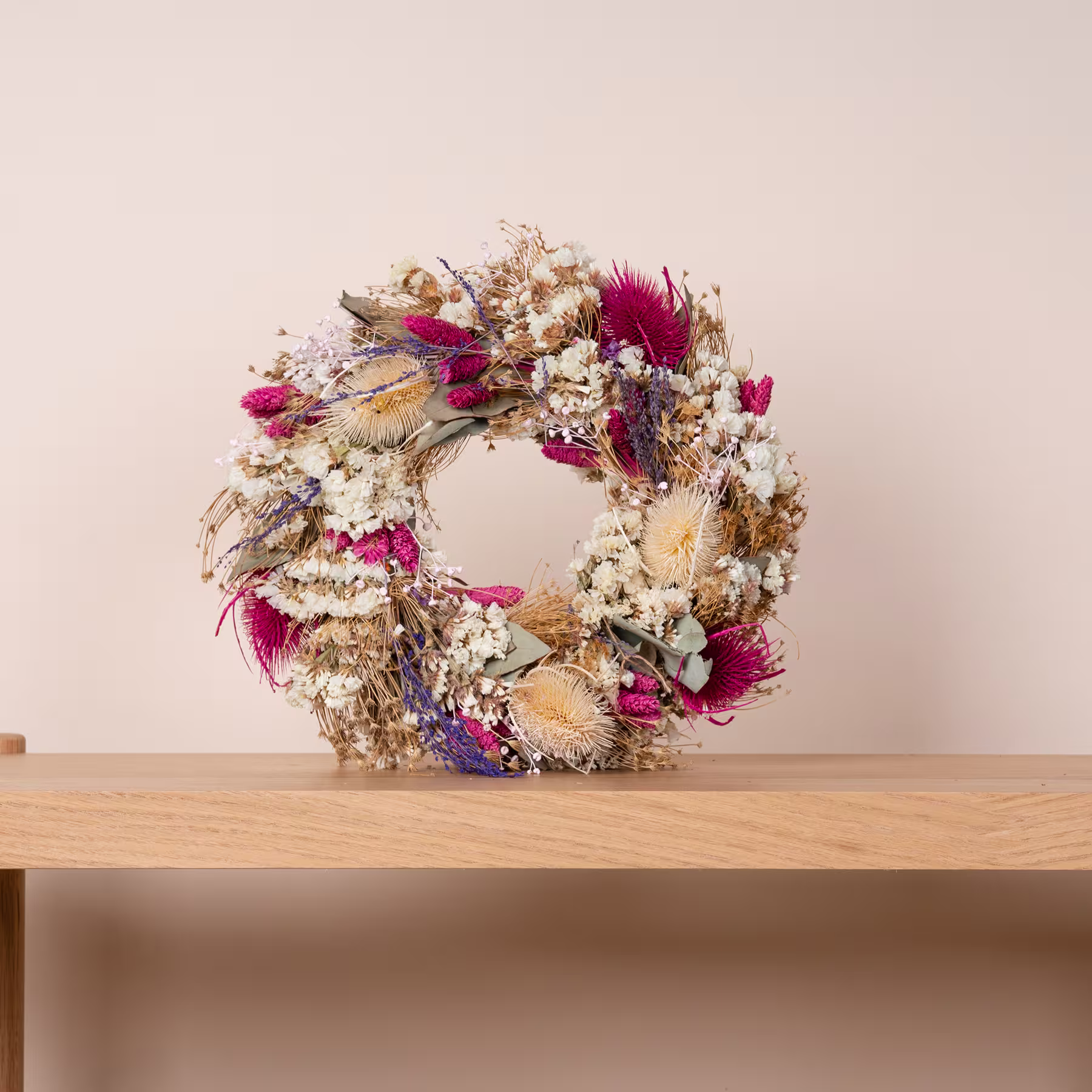 Ein wunderschöner, rustikaler Kranz aus Trockenblumen steht auf einem Holzregal vor einer Wand in neutraler Farbe. (Beschreibungstext KI-generiert)