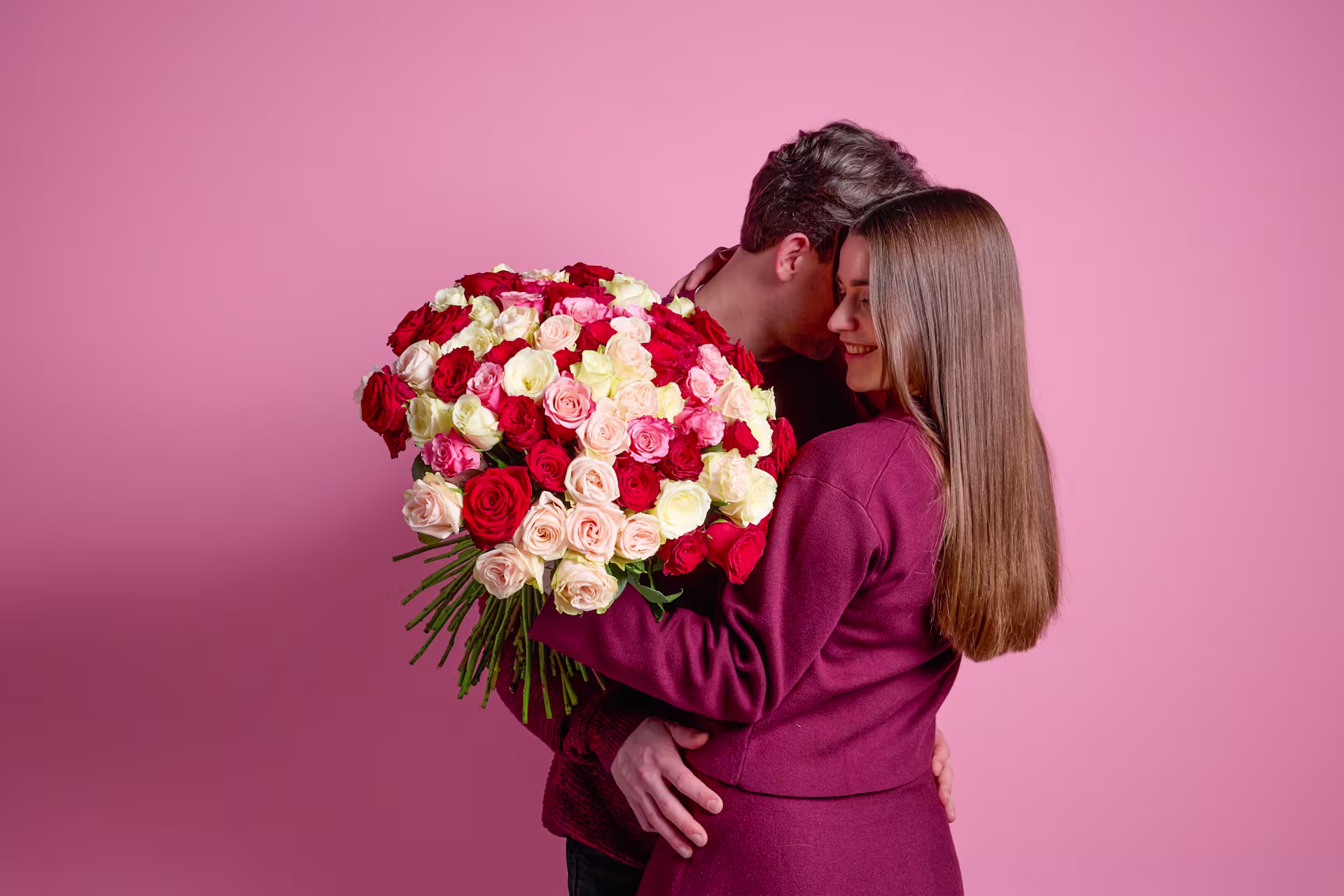 Eine Person in einem burgunderfarbenen Outfit hält einen großen Strauß roter und weißer Rosen vor einem leuchtend rosa Hintergrund. (Beschreibungstext KI-generiert)