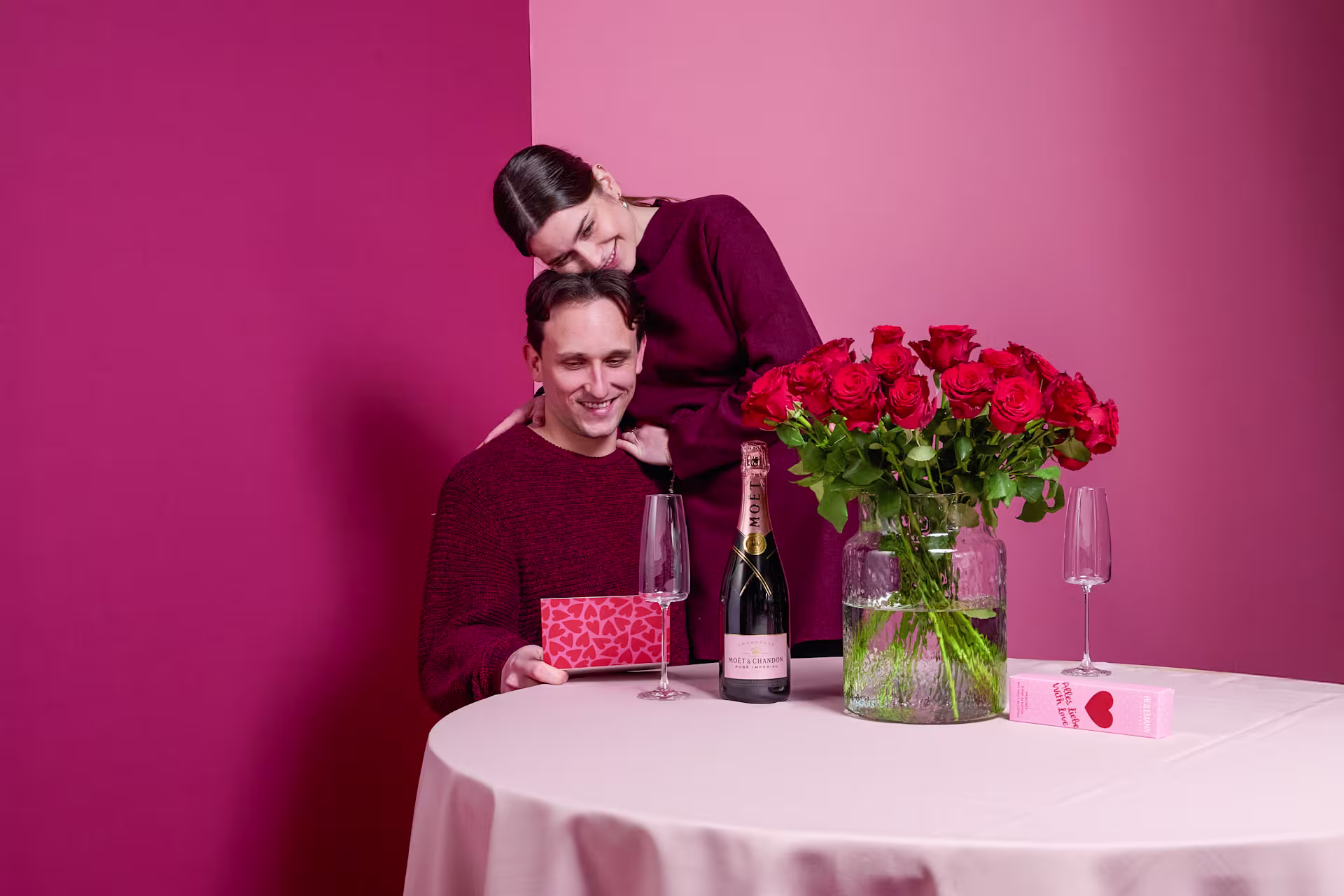 Ein Mann in einem burgunderfarbenen Pullover steht vor einem leuchtend rosa Hintergrund und hält einen Strauß roter Rosen und eine Flasche Wein in der Hand. Auf dem Tisch vor ihm stehen zwei Weingläser. (Beschreibungstext KI-generiert)