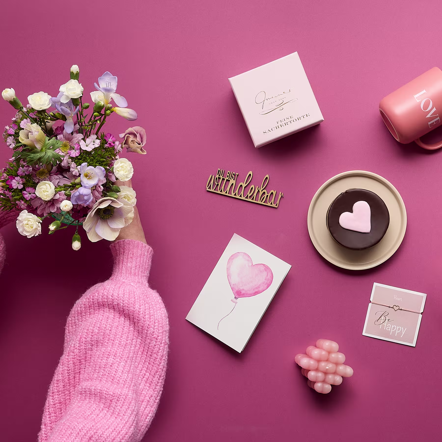 Ein Strauß bunter Blumen, ein rosa Pullover, eine herzförmige Schokolade und verschiedene andere romantische Gegenstände sind auf einem leuchtend rosa Hintergrund arrangiert. (Beschreibungstext KI-generiert)