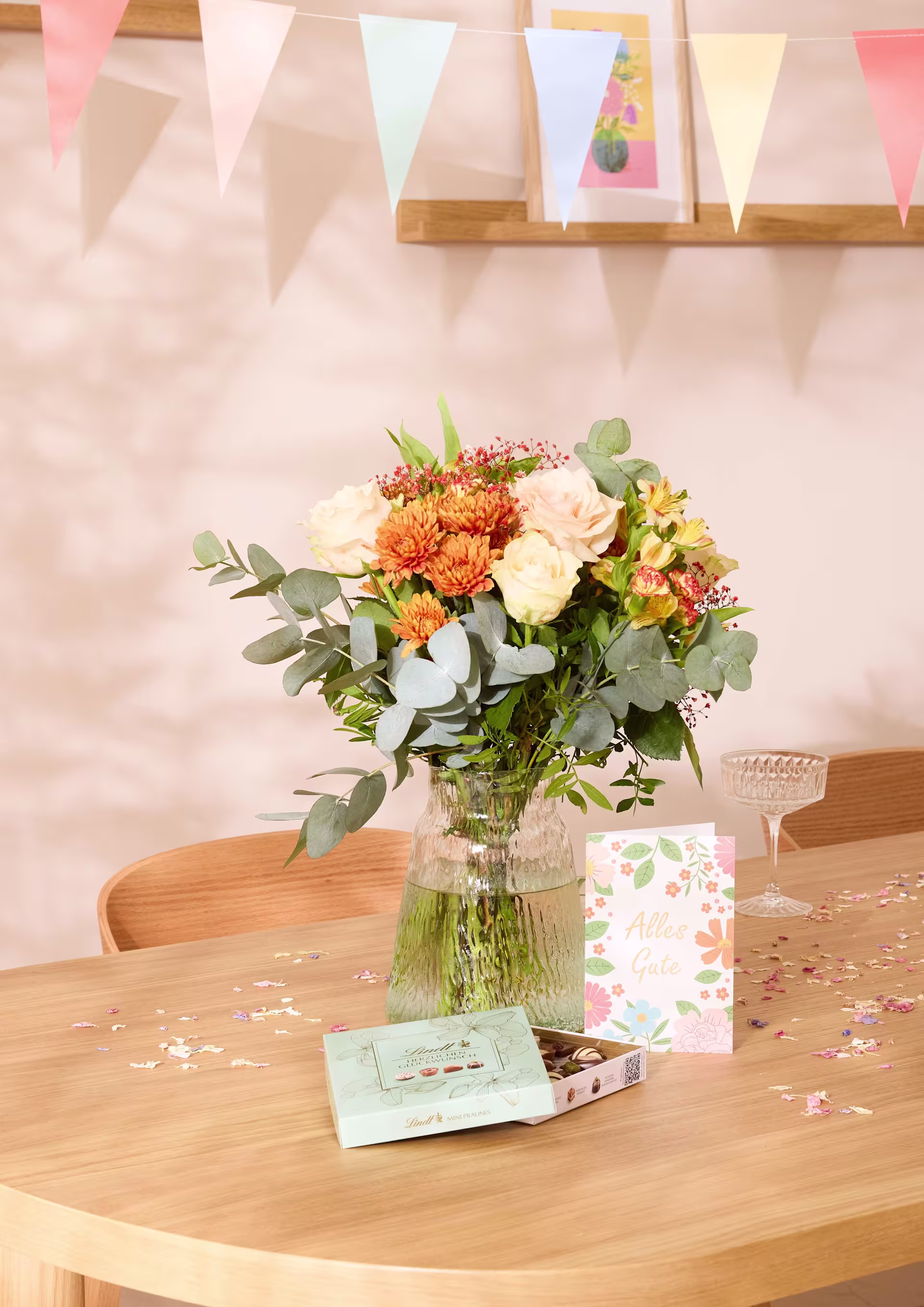 Ein bunter Blumenstrauß steht auf einem Holztisch, umgeben von festlicher Dekoration und Backwaren, und sorgt für eine fröhliche und festliche Atmosphäre. (Beschreibungstext KI-generiert)