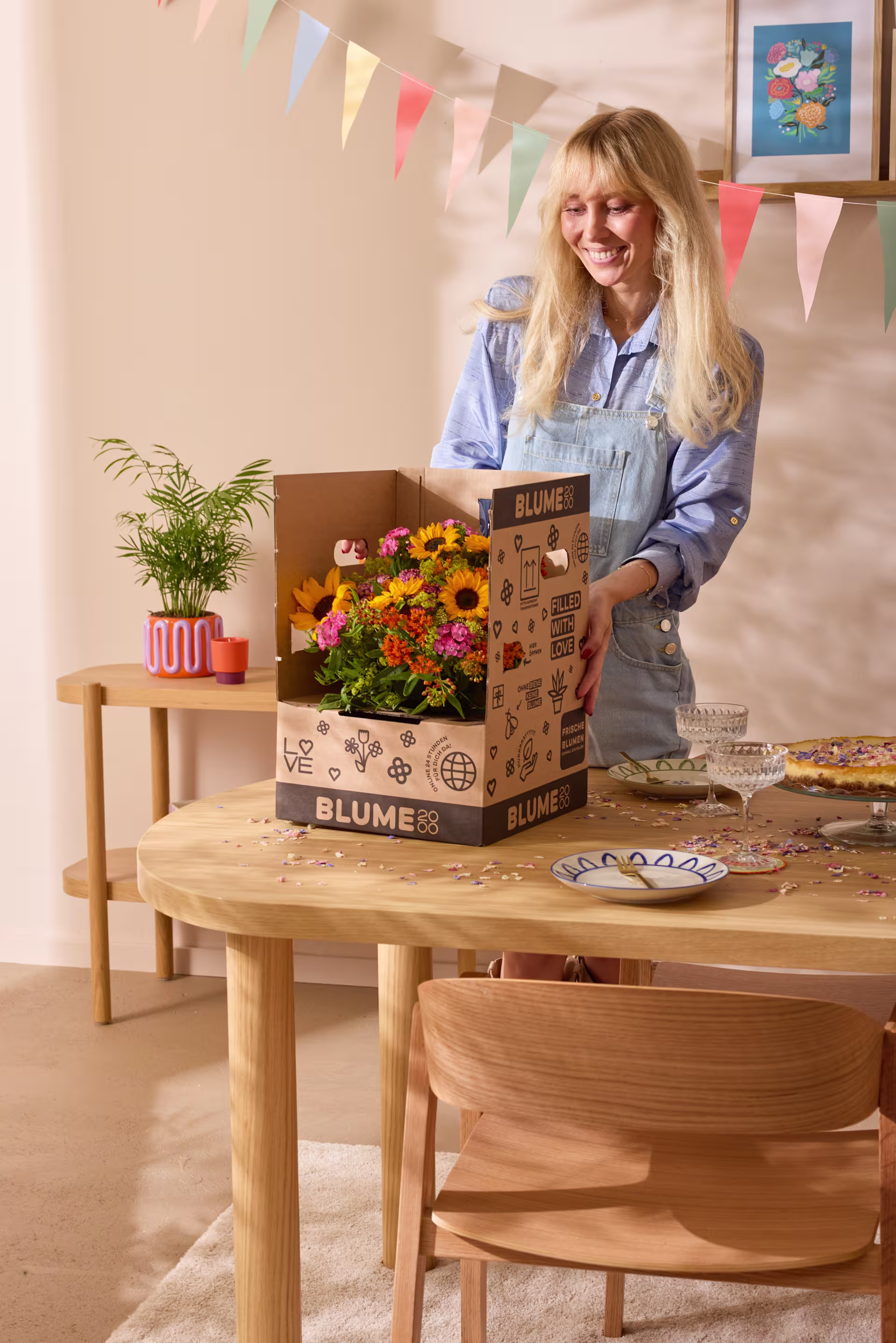 Eine lächelnde Frau mit einer Schachtel bunter Blumen steht in einem gemütlich dekorierten Raum mit einem Holztisch und Stühlen. (Beschreibungstext KI-generiert)