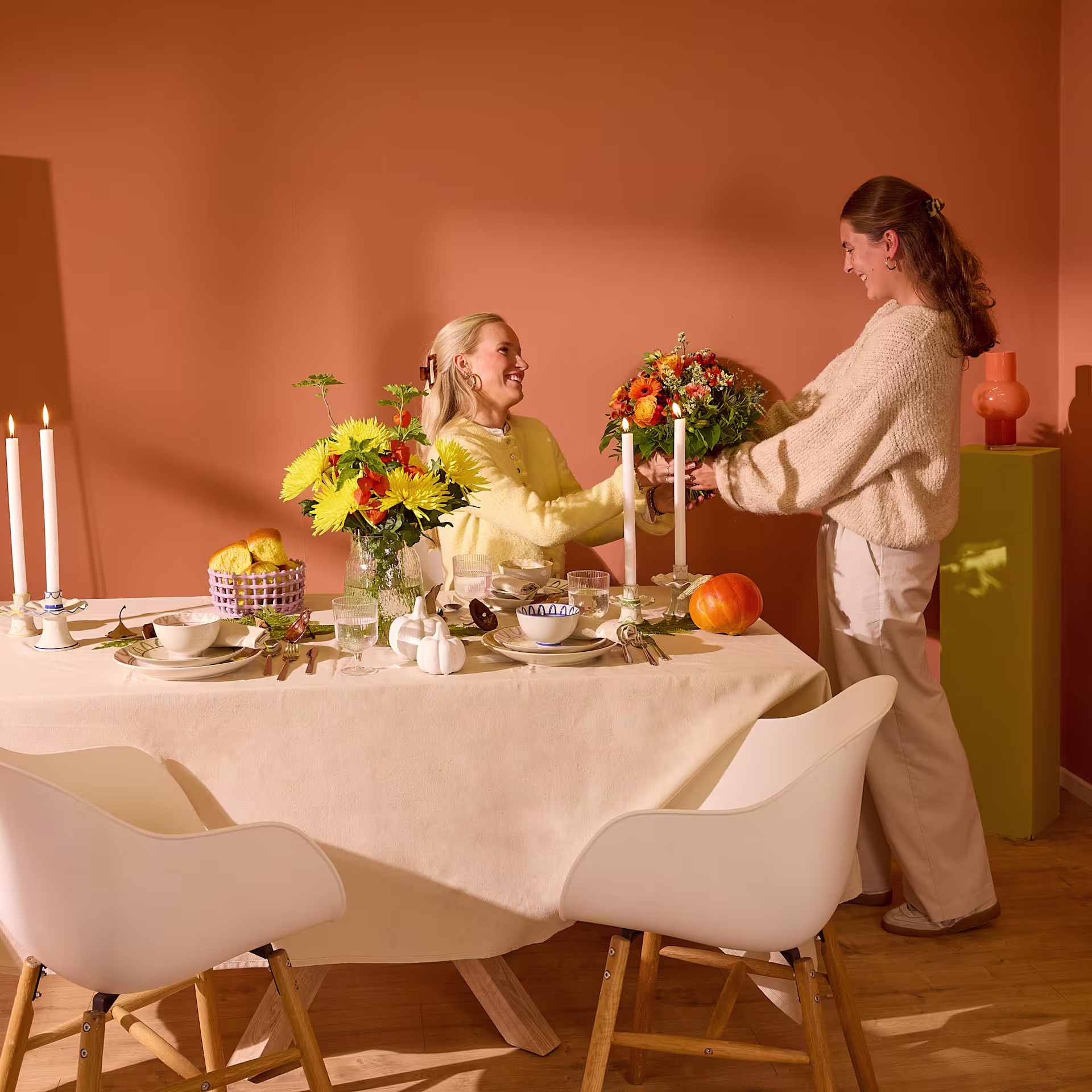 Das Bild zeigt eine gemütliche Essensszene mit einem für eine Mahlzeit gedeckten Tisch. Eine Frau in einem weißen Pullover arrangiert einen Blumenstrauß, während eine andere Frau in einem gelben Kleid am Tisch sitzt. (Beschreibungstext KI-generiert)