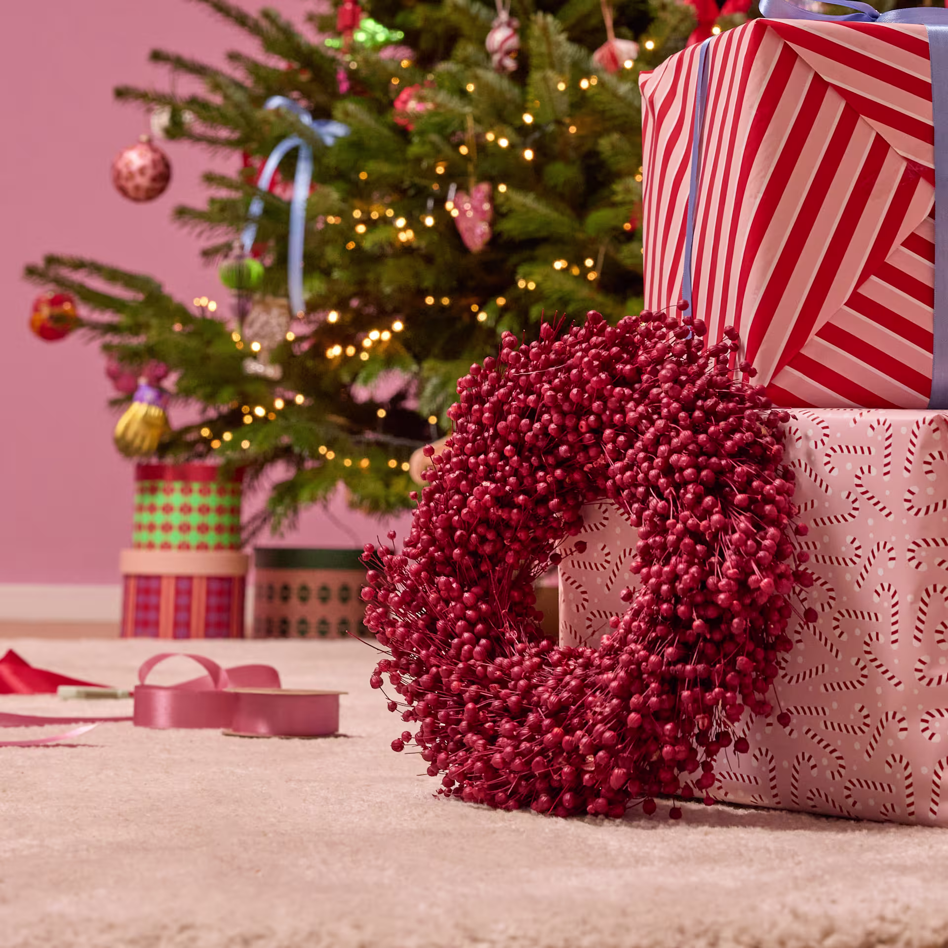 Ein festlicher roter Kranz ziert den Vordergrund, umgeben von einem geschmückten Weihnachtsbaum, verpackten Geschenken und anderen Weihnachtsdekorationen im Hintergrund. (Beschreibungstext KI-generiert)