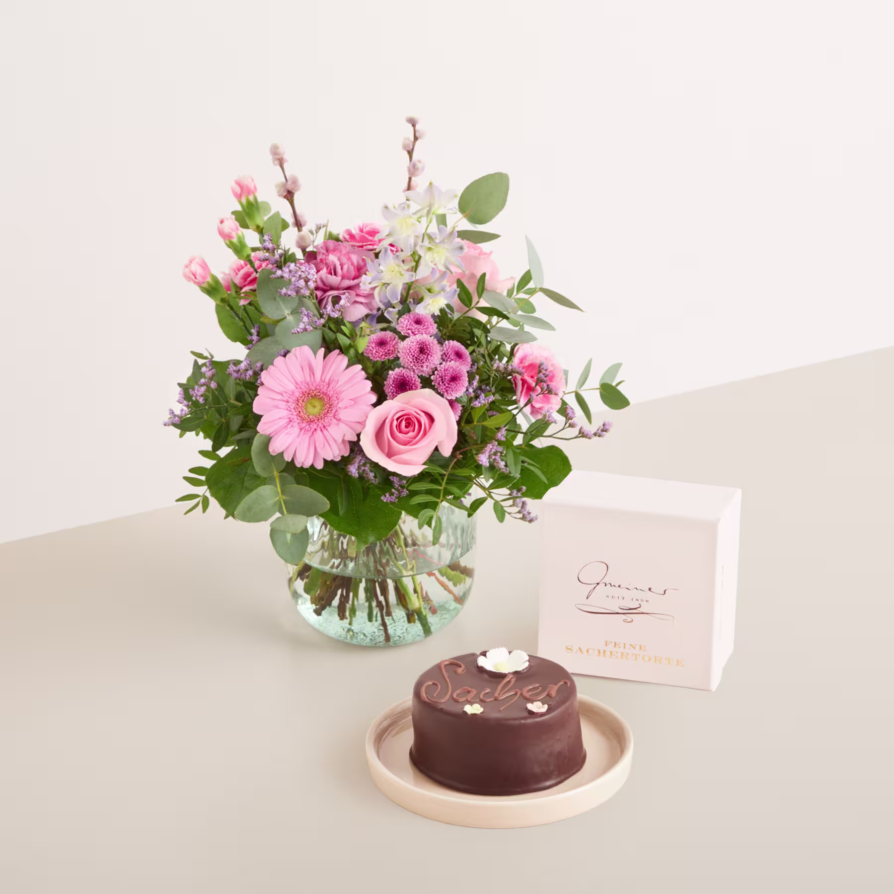 Ein wunderschöner Strauß rosa und weißer Blumen, darunter Rosen und Gerbera, steht in einer Glasvase auf einem Holztisch, dazu ein kleiner Schokoladenkuchen und eine handgeschriebene Notiz. (Beschreibungstext KI-generiert)