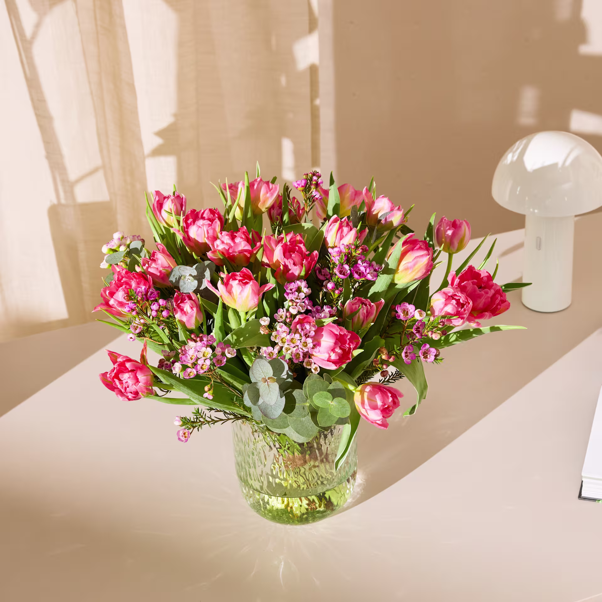 Ein leuchtender Strauß rosa Tulpen und anderer Blumen steht in einer grünen Glasvase auf einem Tisch vor einem weichen, verschwommenen Hintergrund. (Beschreibungstext KI-generiert)
