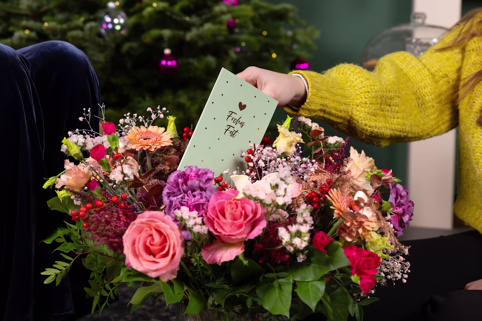 Ein farbenfroher Blumenstrauß mit Rosen, Gänseblümchen und anderen Blüten wird von einer Person in einem gelben Pullover vor dem Hintergrund eines mit funkelnden Lichtern geschmückten Weihnachtsbaums gehalten. (Beschreibungstext KI-generiert)
