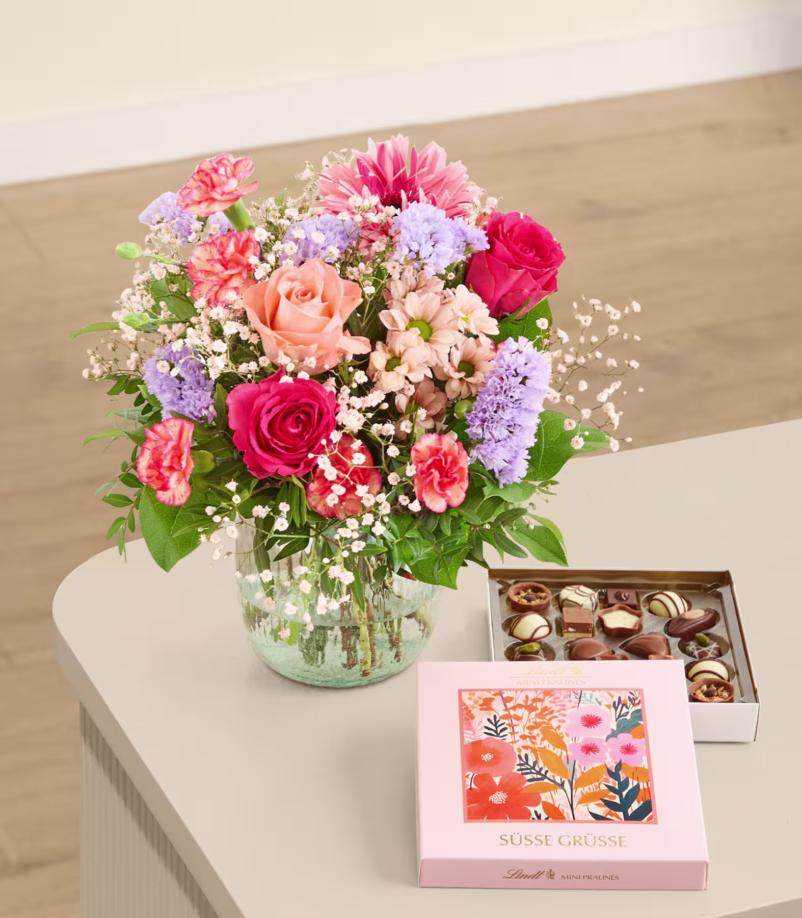 Ein farbenfroher Strauß aus Rosen, Nelken und Gänseblümchen steht in einer Glasvase auf einer hellen Unterlage, daneben eine Schachtel Pralinen. (Beschreibungstext KI-generiert)