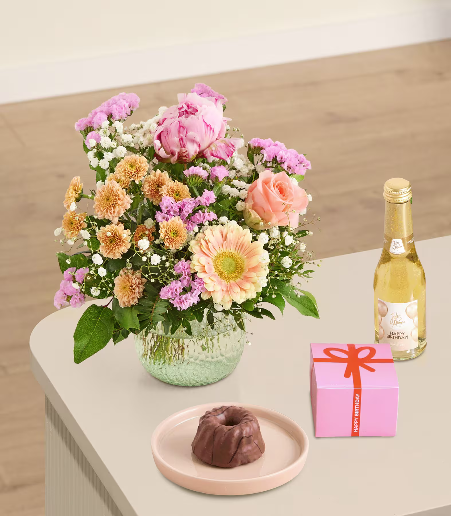 Auf einem Holztisch steht ein prächtiger Strauß aus rosa Pfingstrosen, gelben Gänseblümchen und anderen farbenfrohen Blumen, dazu eine Flasche Wein und eine kleine Geschenkbox. (Beschreibungstext KI-generiert)