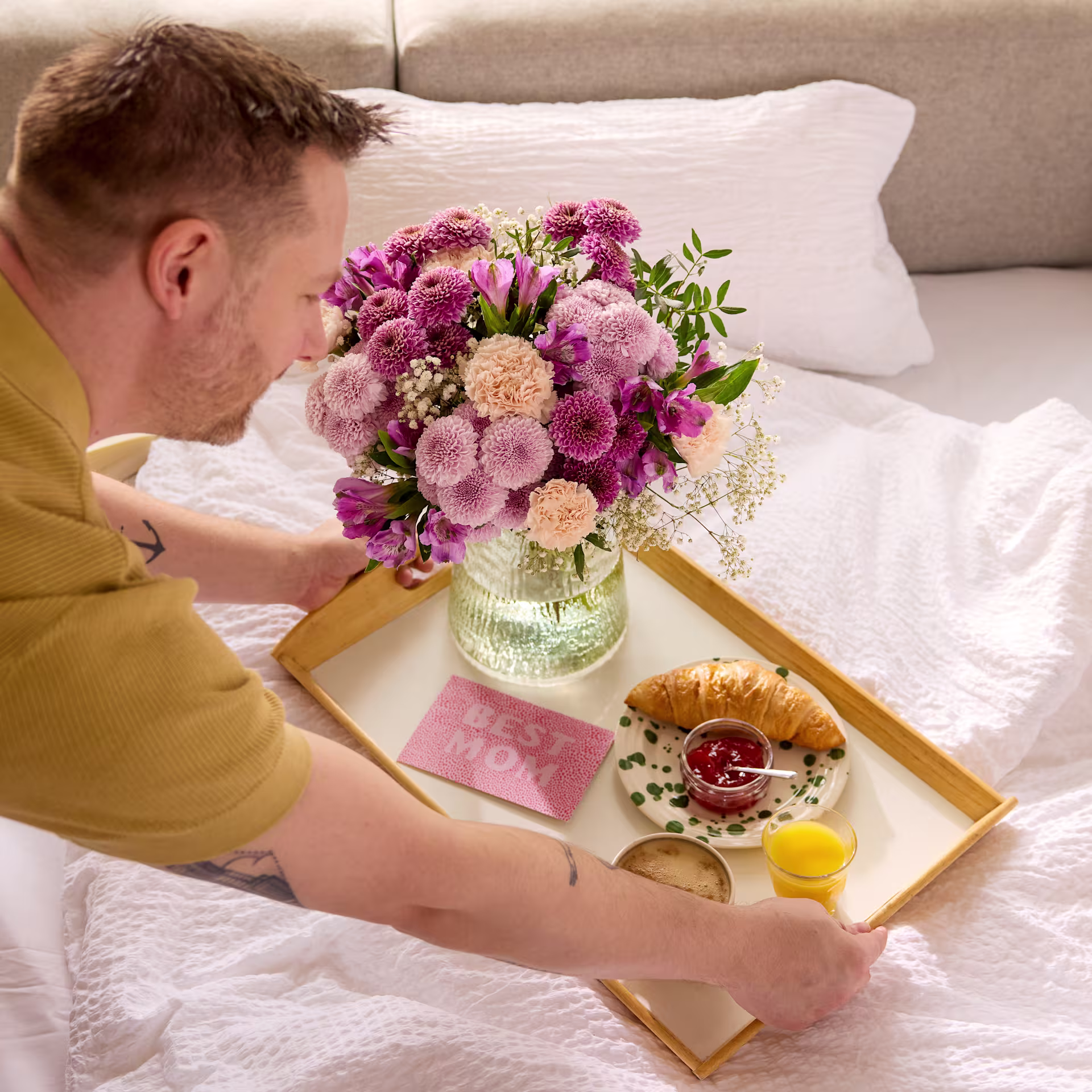 Ein Mann in einem gelben Hemd hält ein Tablett mit einer Vase voller bunter Blumen, einem Croissant und anderen Frühstückszutaten auf einem Bett mit weißer Bettwäsche. (Beschreibungstext KI-generiert)