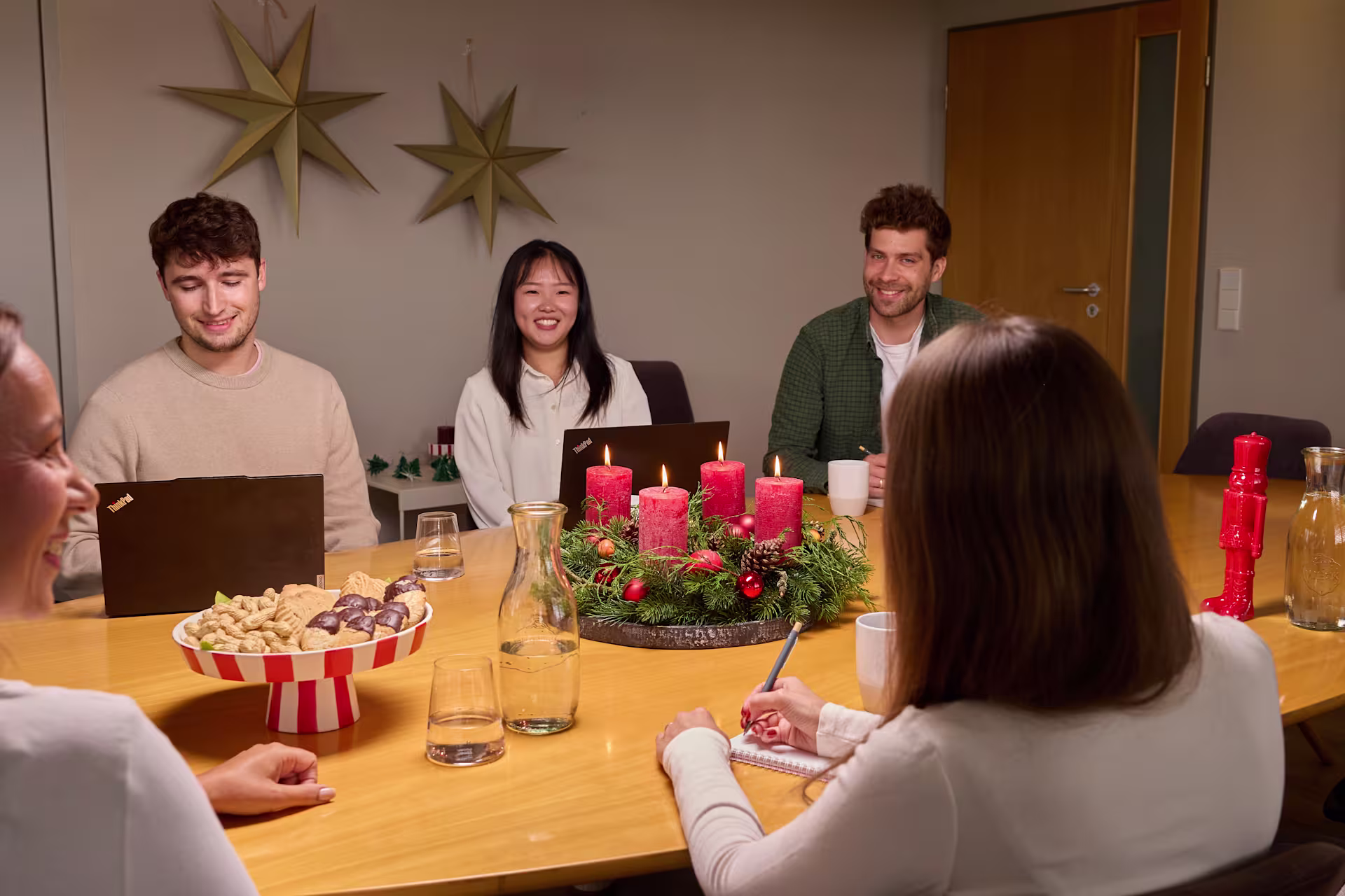 Vier Personen sitzen um einen Holztisch mit roten Kerzen und Snacks im Vordergrund, vor einer schlichten Wand, die mit zwei großen goldenen Sternen verziert ist. (Beschreibungstext KI-generiert)