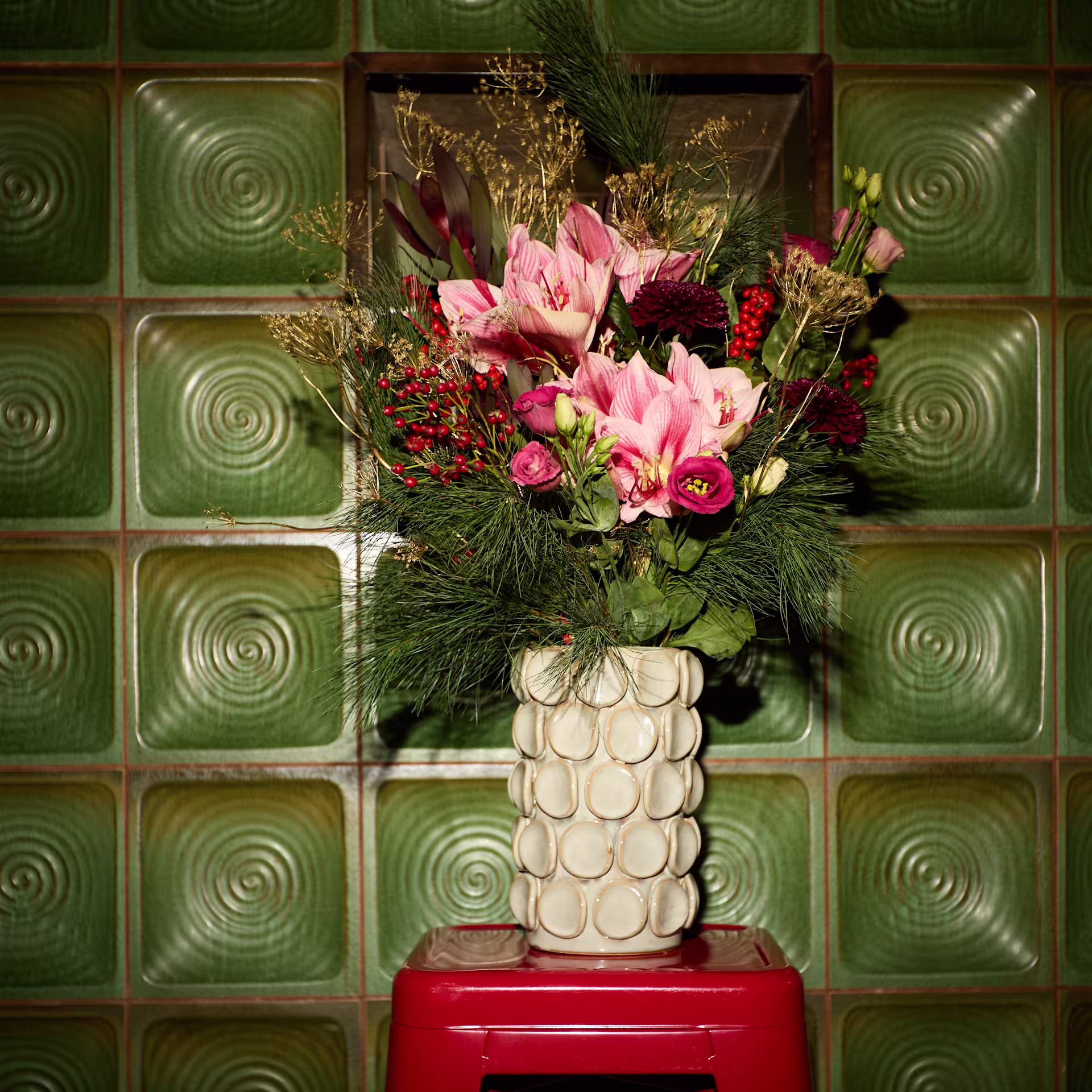 Ein farbenfrohes Blumenarrangement in einer strukturierten weißen Vase steht auf einem roten Hocker vor einem Hintergrund aus grünen, spiralförmig gemusterten Fliesen. (Beschreibungstext KI-generiert)