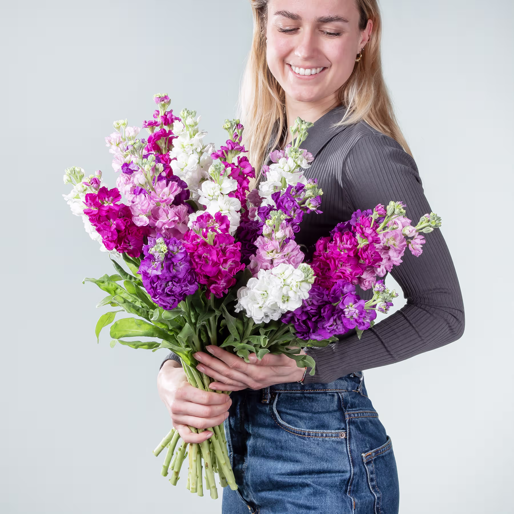 Eine lächelnde junge Frau hält einen großen Strauß leuchtend rosa, violetter und weißer Blumen vor einem schlichten hellen Hintergrund. (Beschreibungstext KI-generiert)