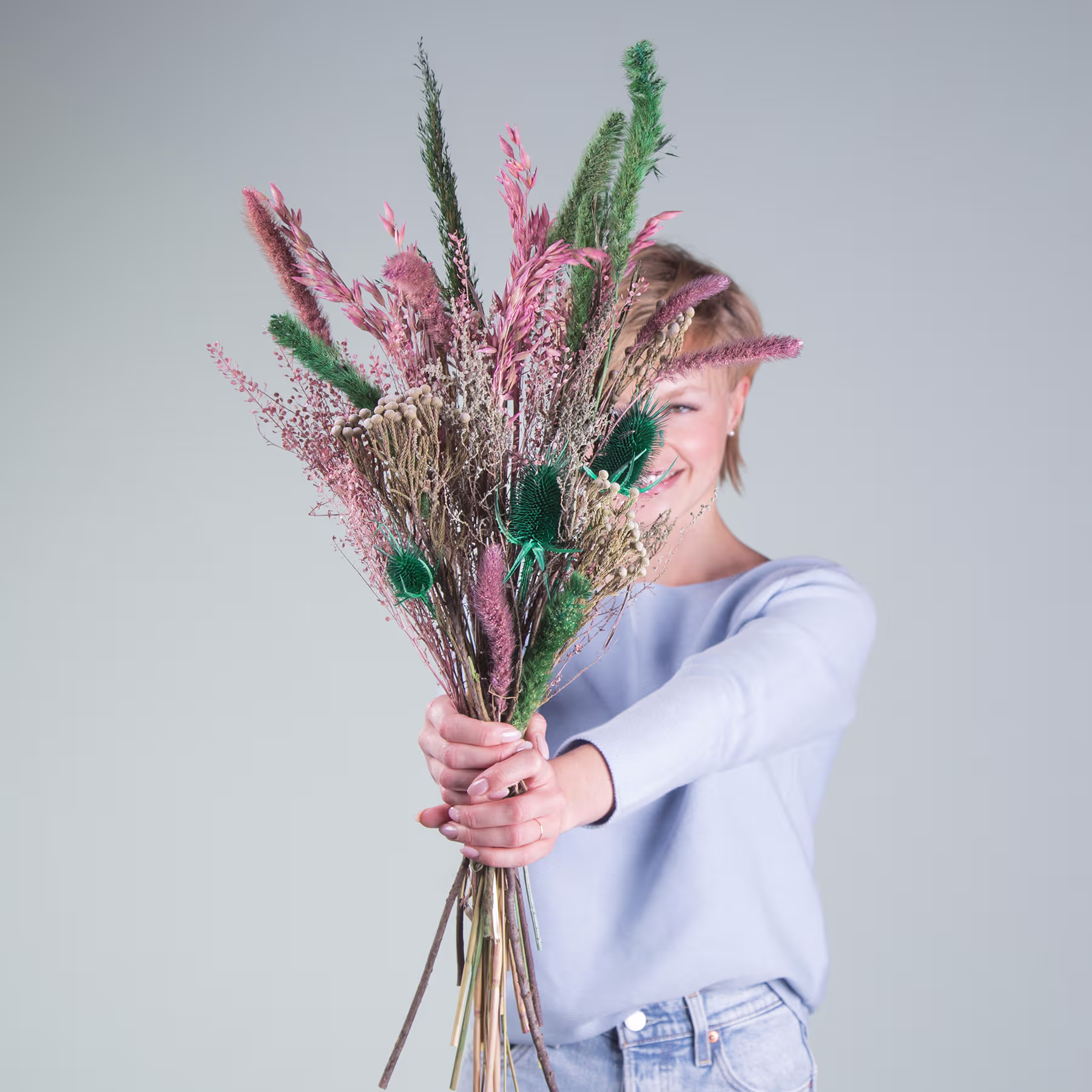 Eine Person hält einen großen Strauß lebendiger, bunter Trockenblumen und Pflanzen vor einem schlichten, hellen Hintergrund. (Beschreibungstext KI-generiert)