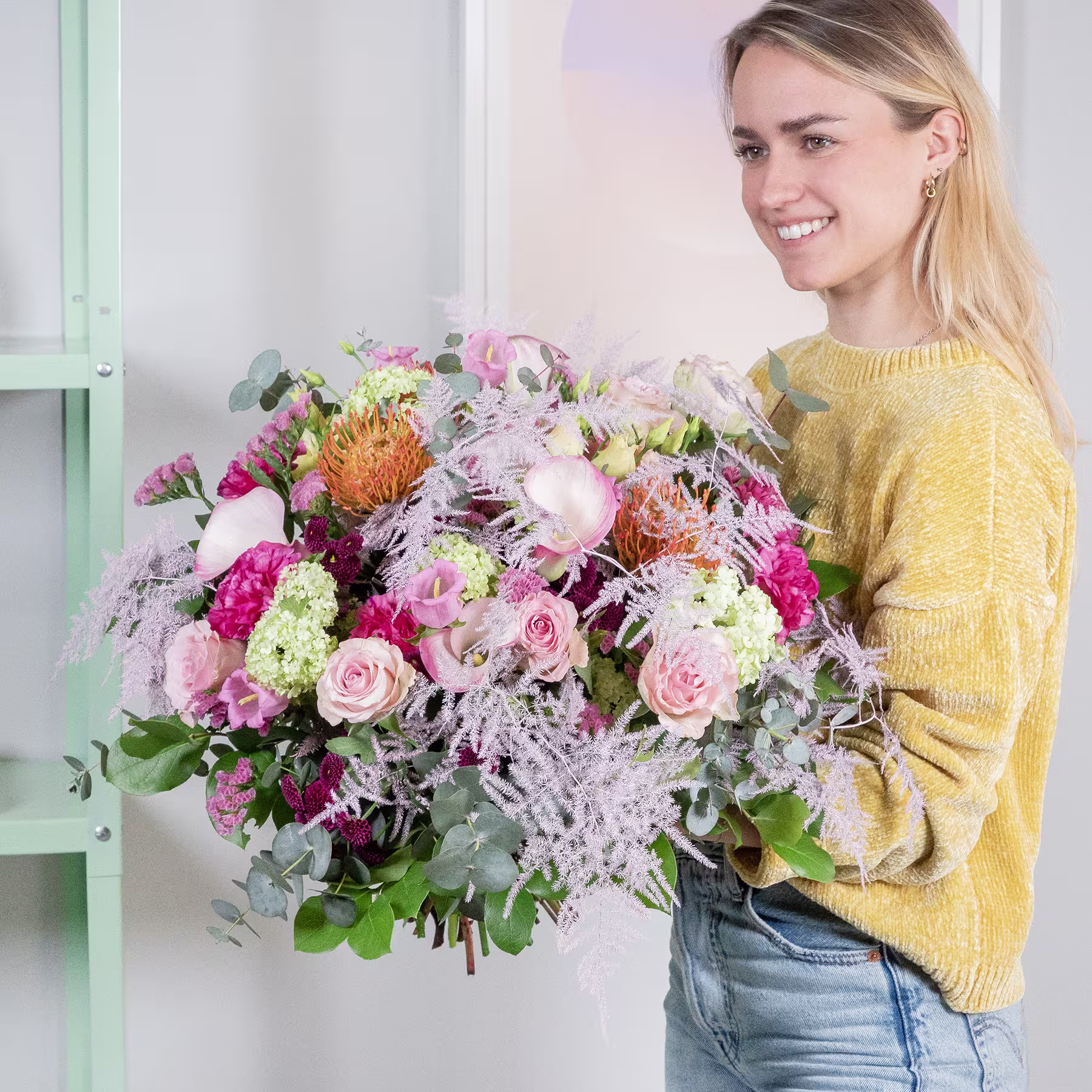 Eine lächelnde junge Frau in einem gelben Pullover hält einen großen, leuchtenden Strauß bunter Blumen vor einem hellen Hintergrund. (Beschreibungstext KI-generiert)